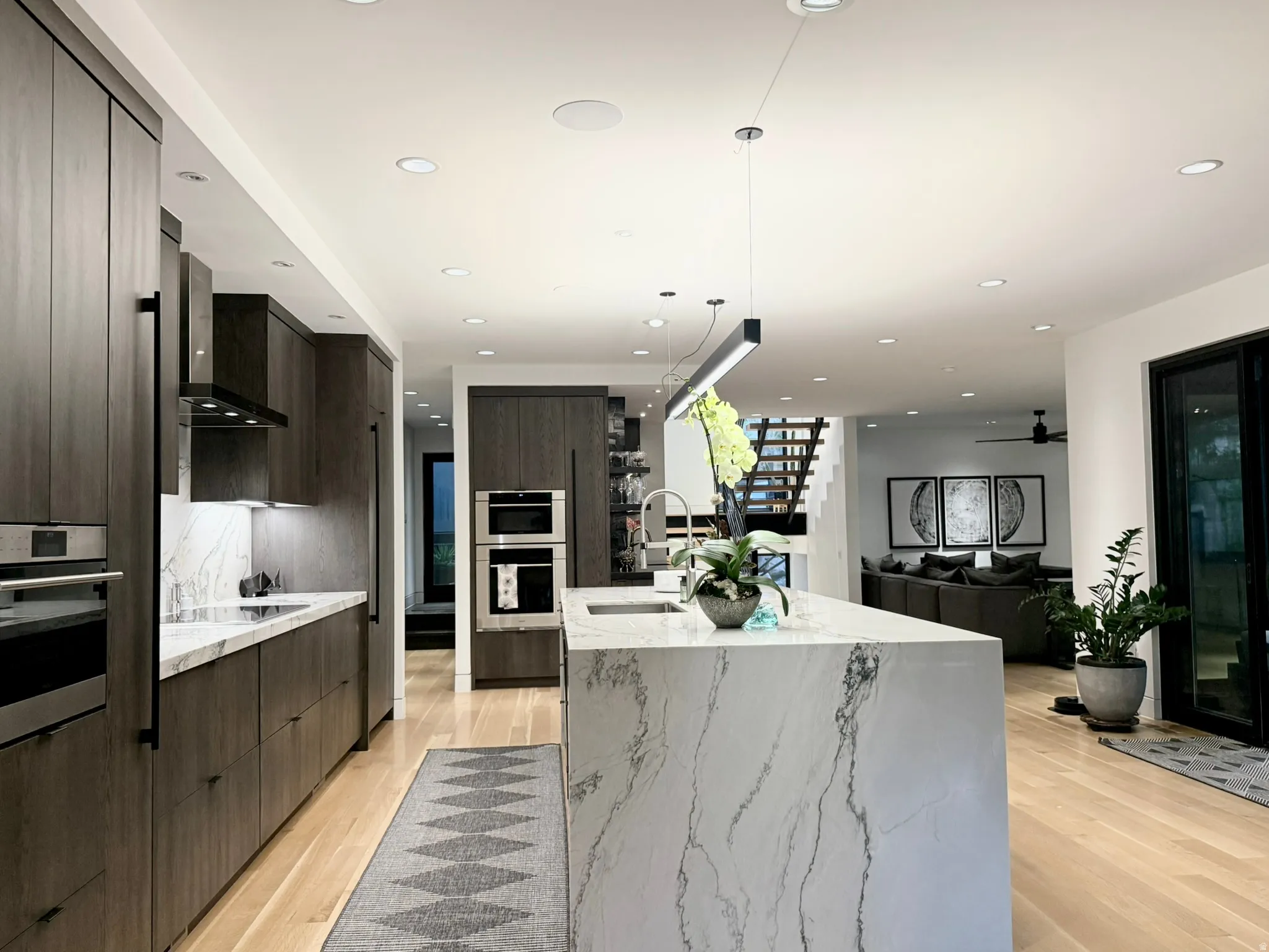 Modern kitchen with a large waterfall island featuring a stone countertop, wood-finish cabinetry, built-in stainless steel appliances, and light wood flooring