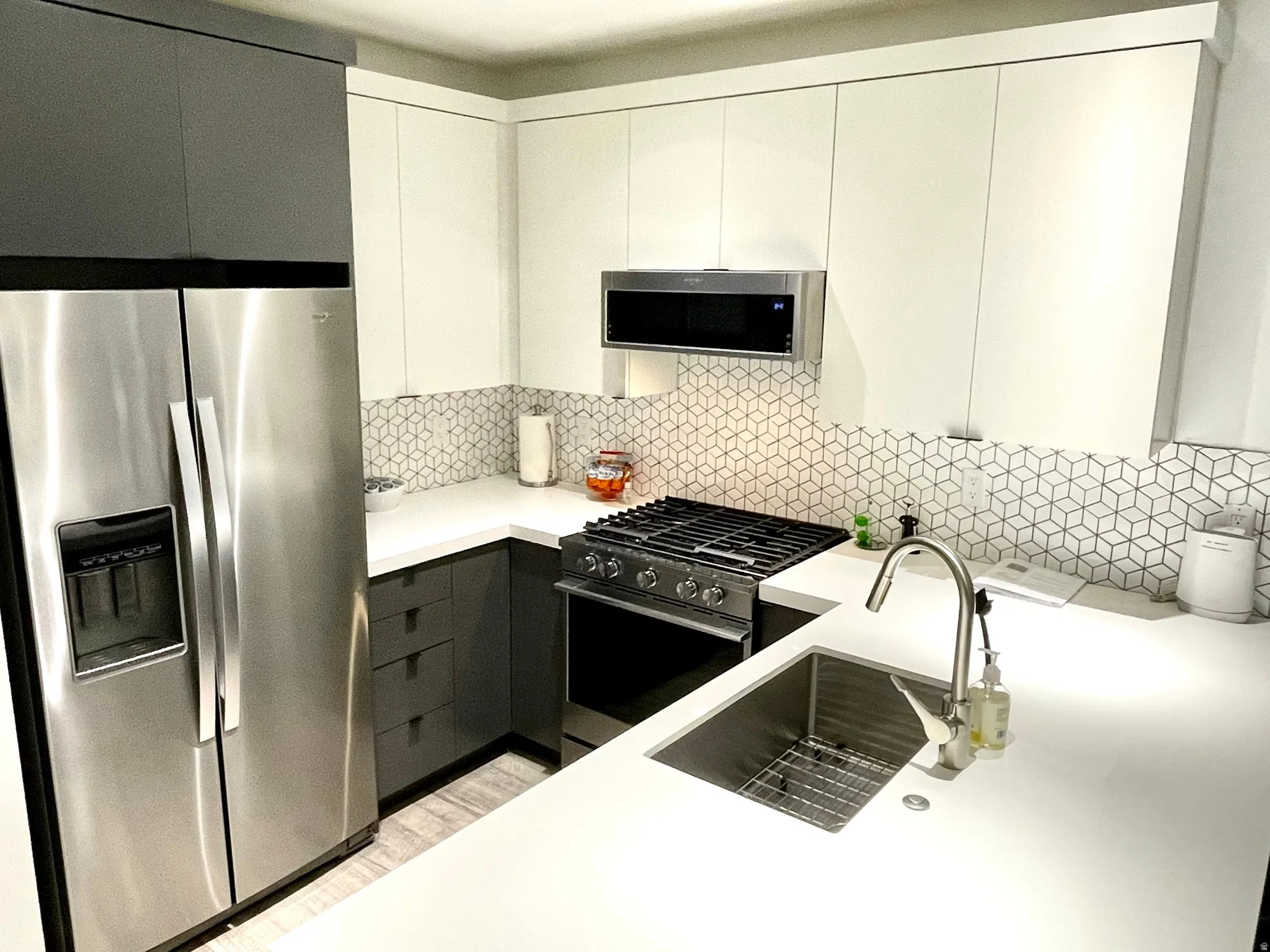 Modern kitchen featuring a stainless steel side-by-side refrigerator, white solid surface countertops, a mosaic tile backsplash with geometric pattern, two-tone cabinetry with white upper and dark lower units, and a stainless steel undermount sink with a