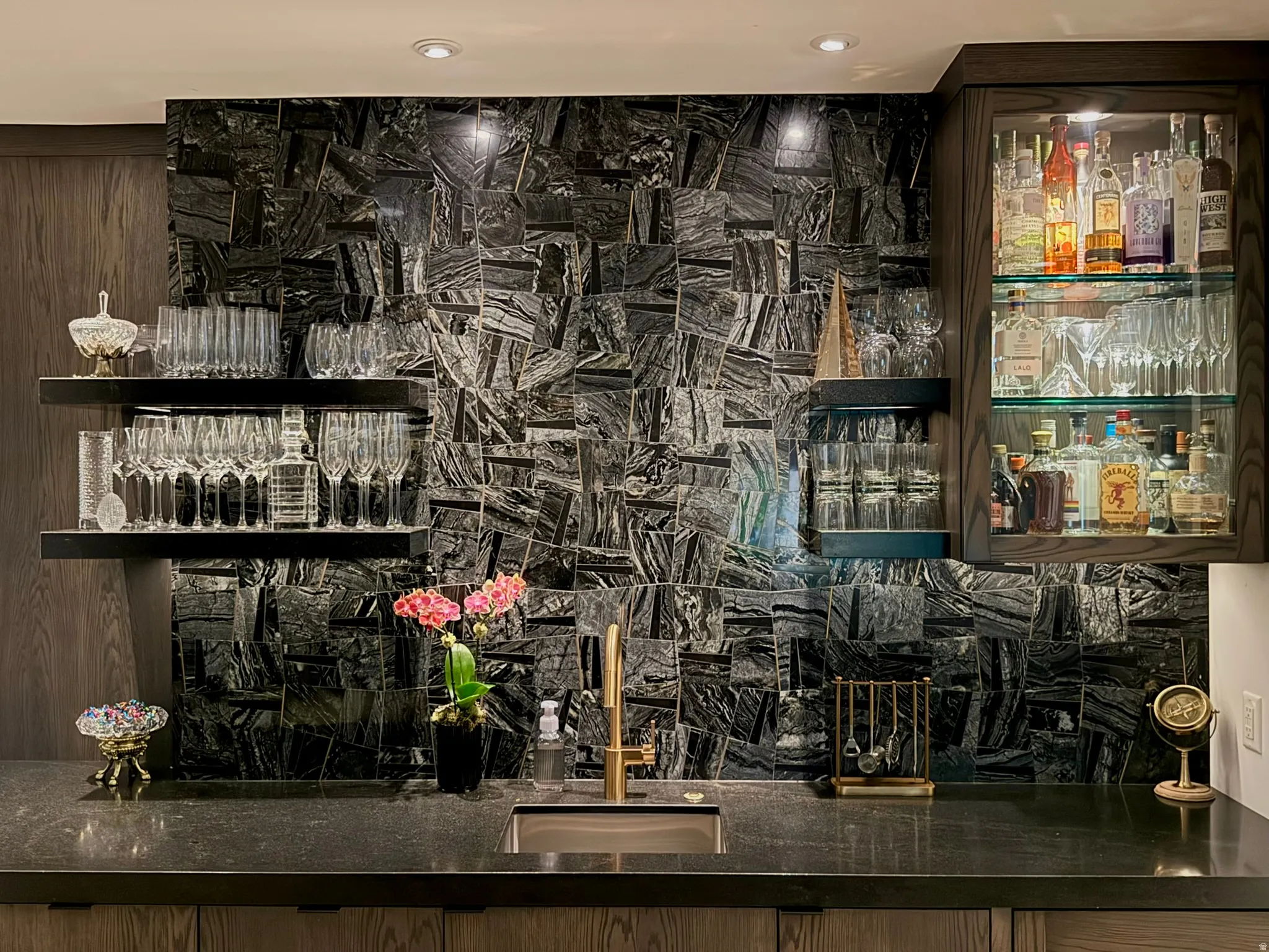 Wet bar featuring a dark stone countertop, an integrated sink with a gold-tone faucet, and a dramatic black stone tile backsplash