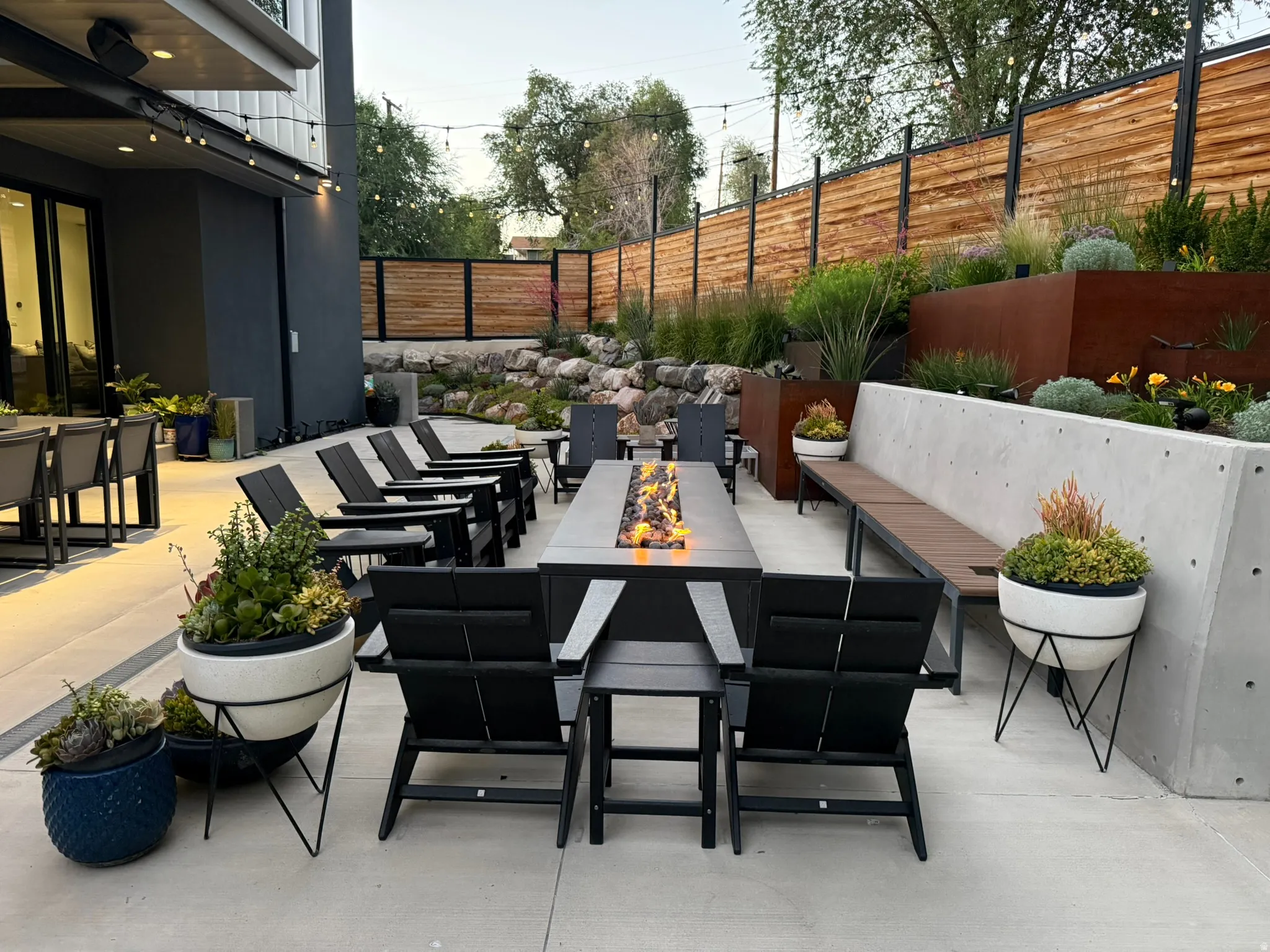 Spacious outdoor patio area featuring a fire pit, concrete hardscaping, tiered landscaping with mature plantings, a modern slatted wood fence, and string lighting