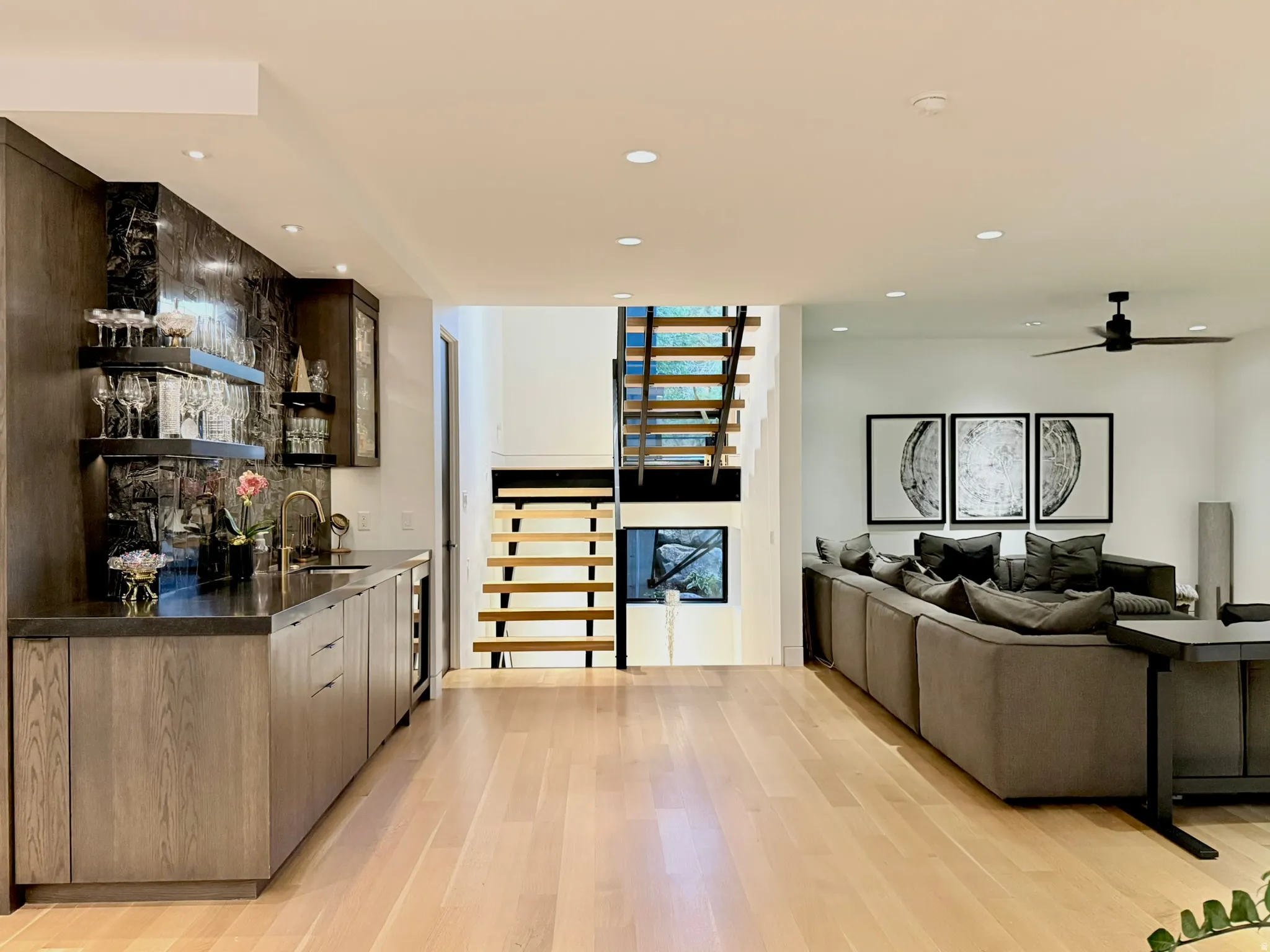 Spacious open-concept area featuring light wood-finish flooring, a built-in wet bar with dark cabinetry and stone-look backsplash, a contemporary open-riser staircase, recessed lighting, and a ceiling fan