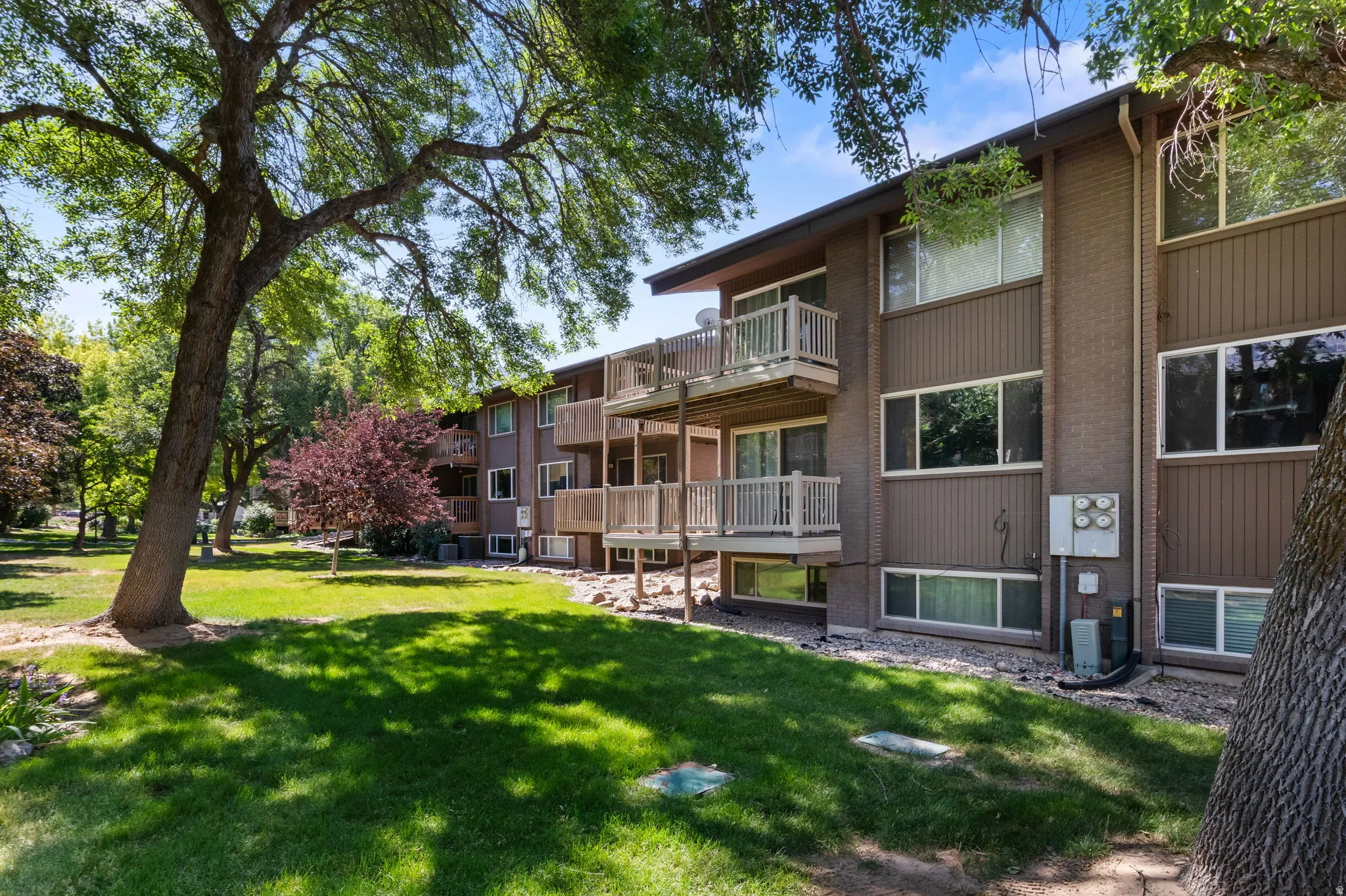 Multi-story building featuring private balconies, brick accents, and a surrounding green space with mature trees