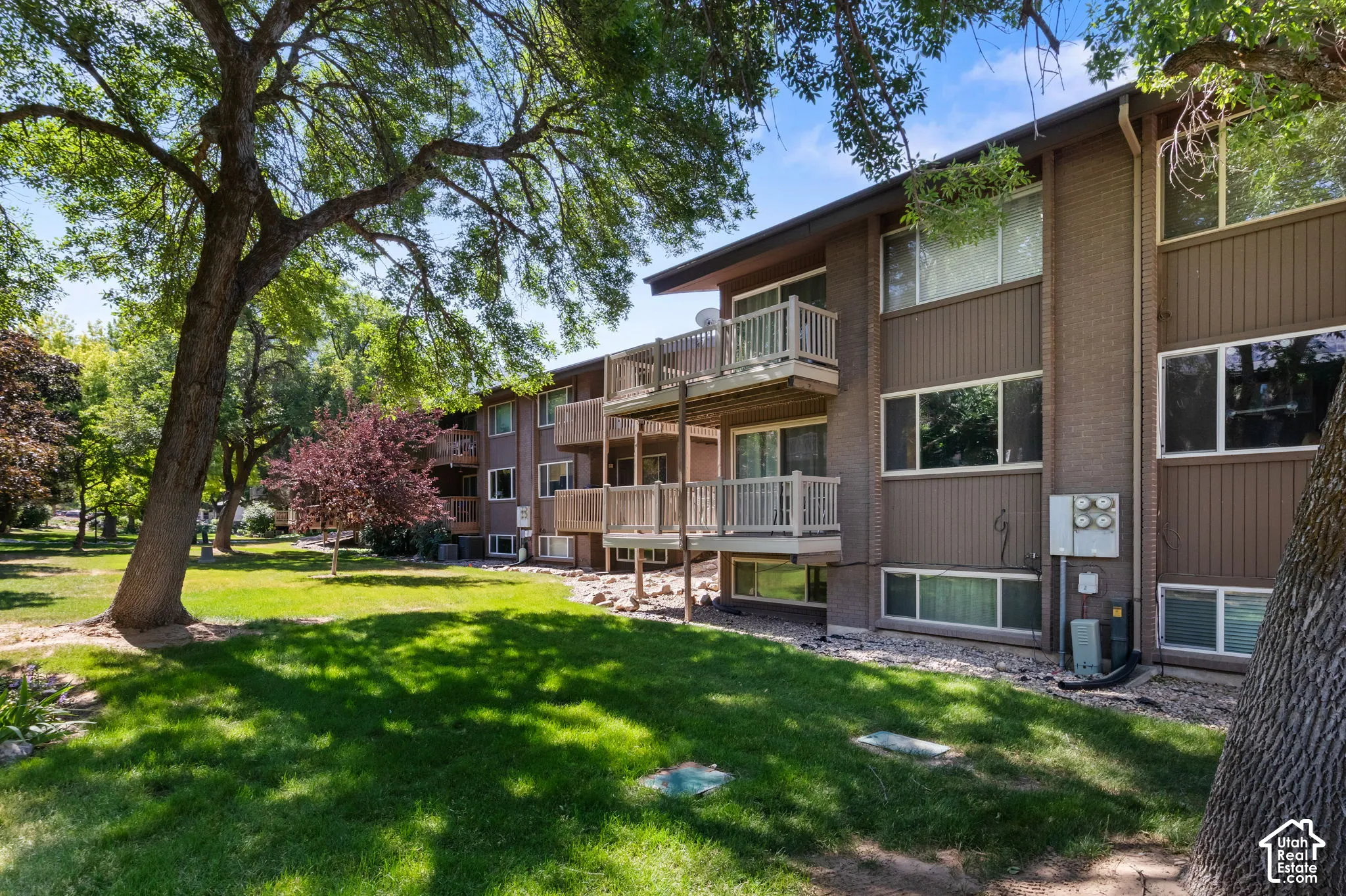 Multi-story building featuring private balconies, brick accents, and a surrounding green space with mature trees
