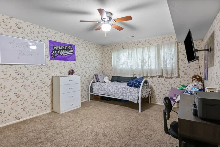 Carpeted room featuring patterned wallpaper, a ceiling fan with light fixture, and window with sheer curtains