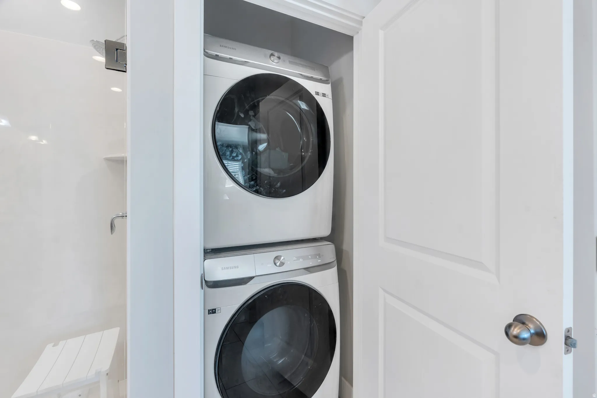 Compact laundry area featuring a stacked washer and dryer, adjacent to a shower with white paneling and a built-in shelf