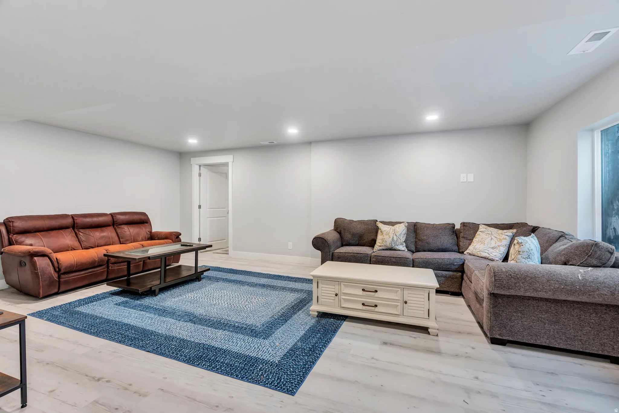 Spacious living area featuring light-toned wood-finish flooring, recessed lighting, neutral wall paint, a white door frame, and a visible window opening