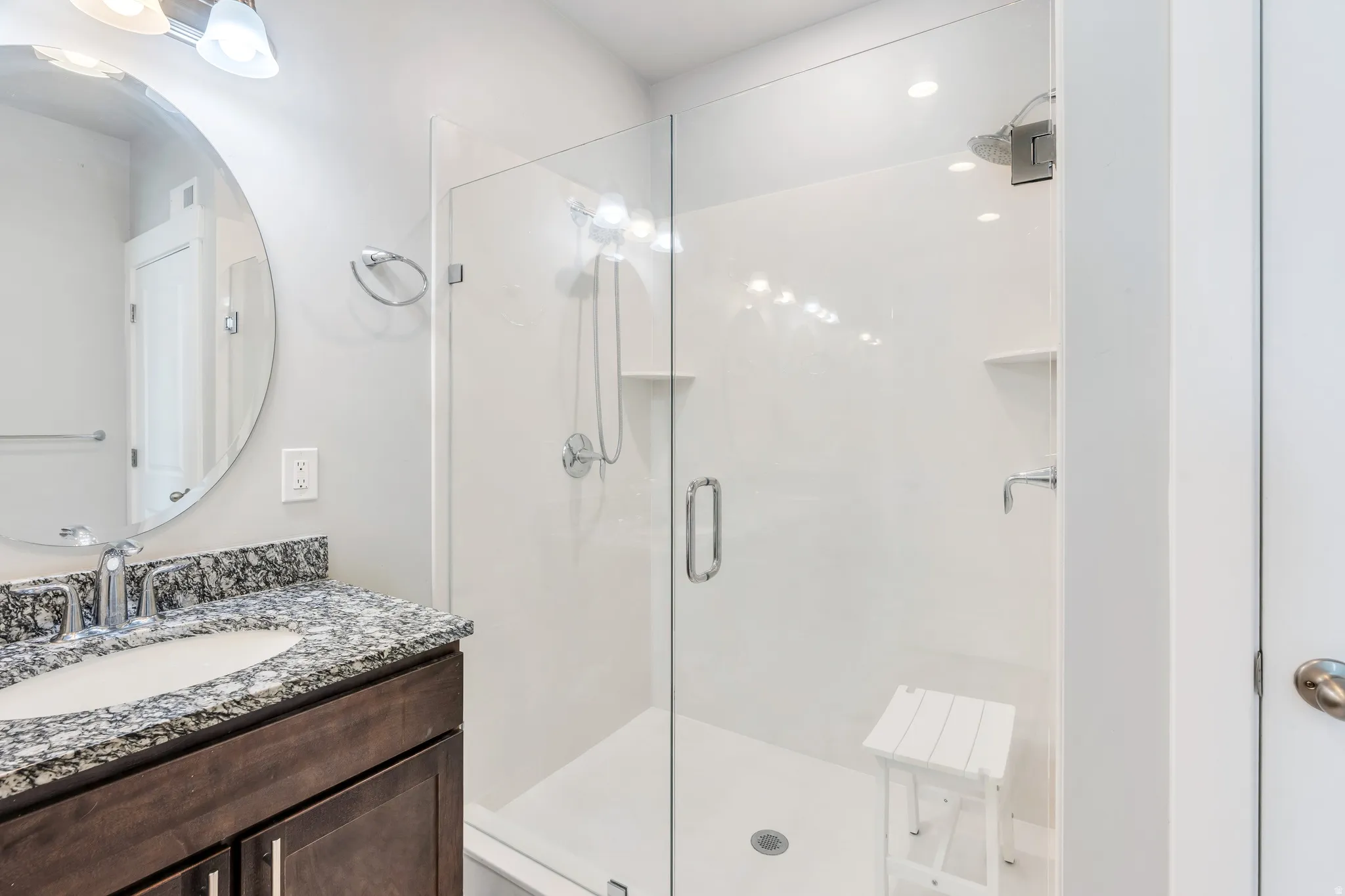 Bathroom featuring a glass-enclosed shower, a vanity with a dark wood-finish cabinet, a speckled countertop, an undermount sink, and a circular mirror