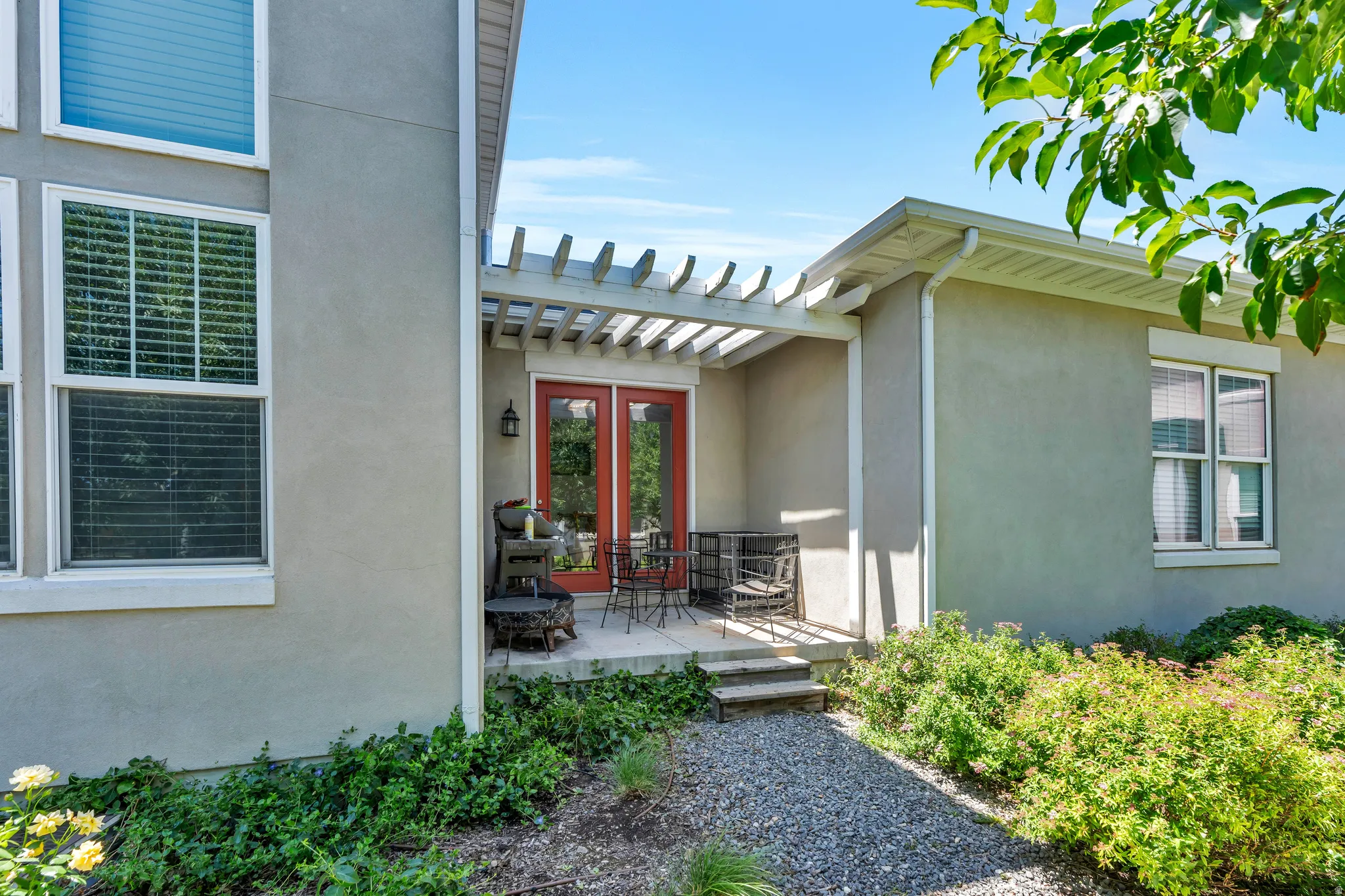 Stucco exterior featuring a pergola-covered patio with red French doors, gravel pathway, and established landscaping