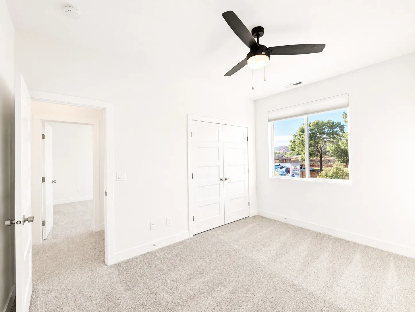 Light-filled room featuring a large window, neutral carpeting, a ceiling fan with integrated lighting, and white bi-fold closet doors