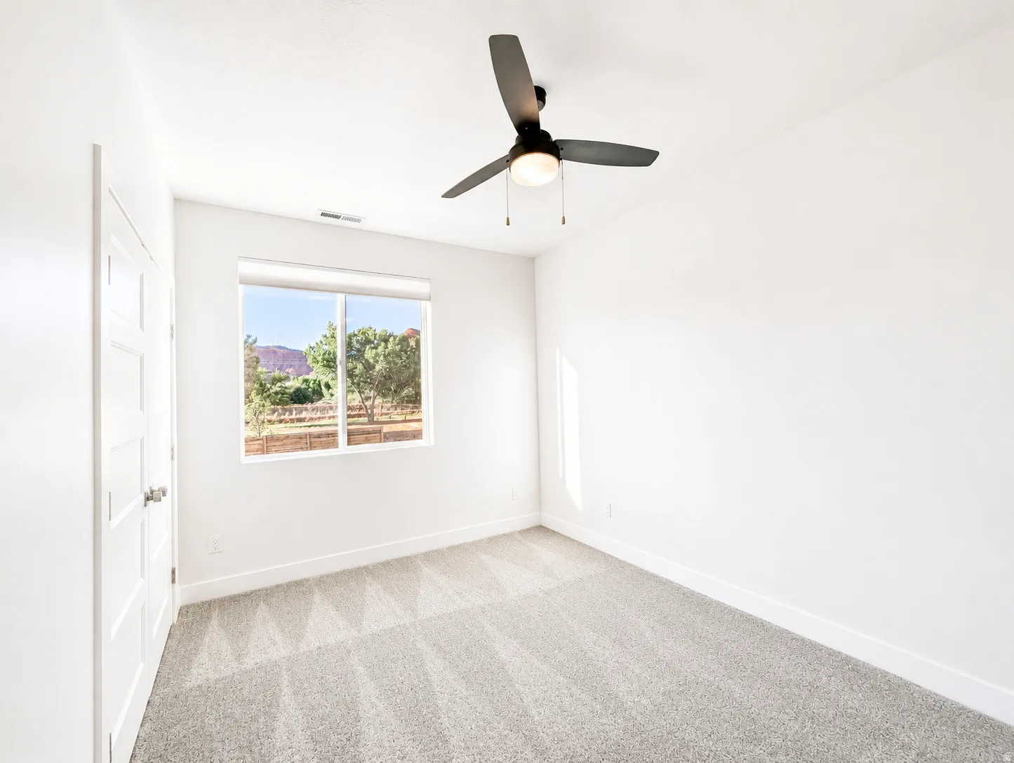 Bright room featuring a large window, neutral carpeting, a ceiling fan, a modern paneled door, and white baseboards