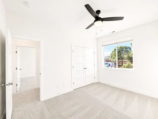 Light-filled room featuring a large window, neutral carpeting, a ceiling fan with integrated lighting, and white bi-fold closet doors