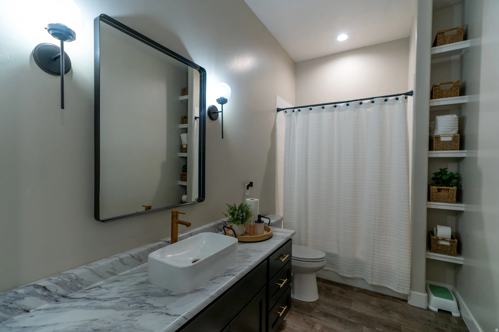 Bathroom featuring a rectangular vessel sink, wood-finish flooring, built-in shelving, a shower with a white curtain, and wall-mounted sconces
