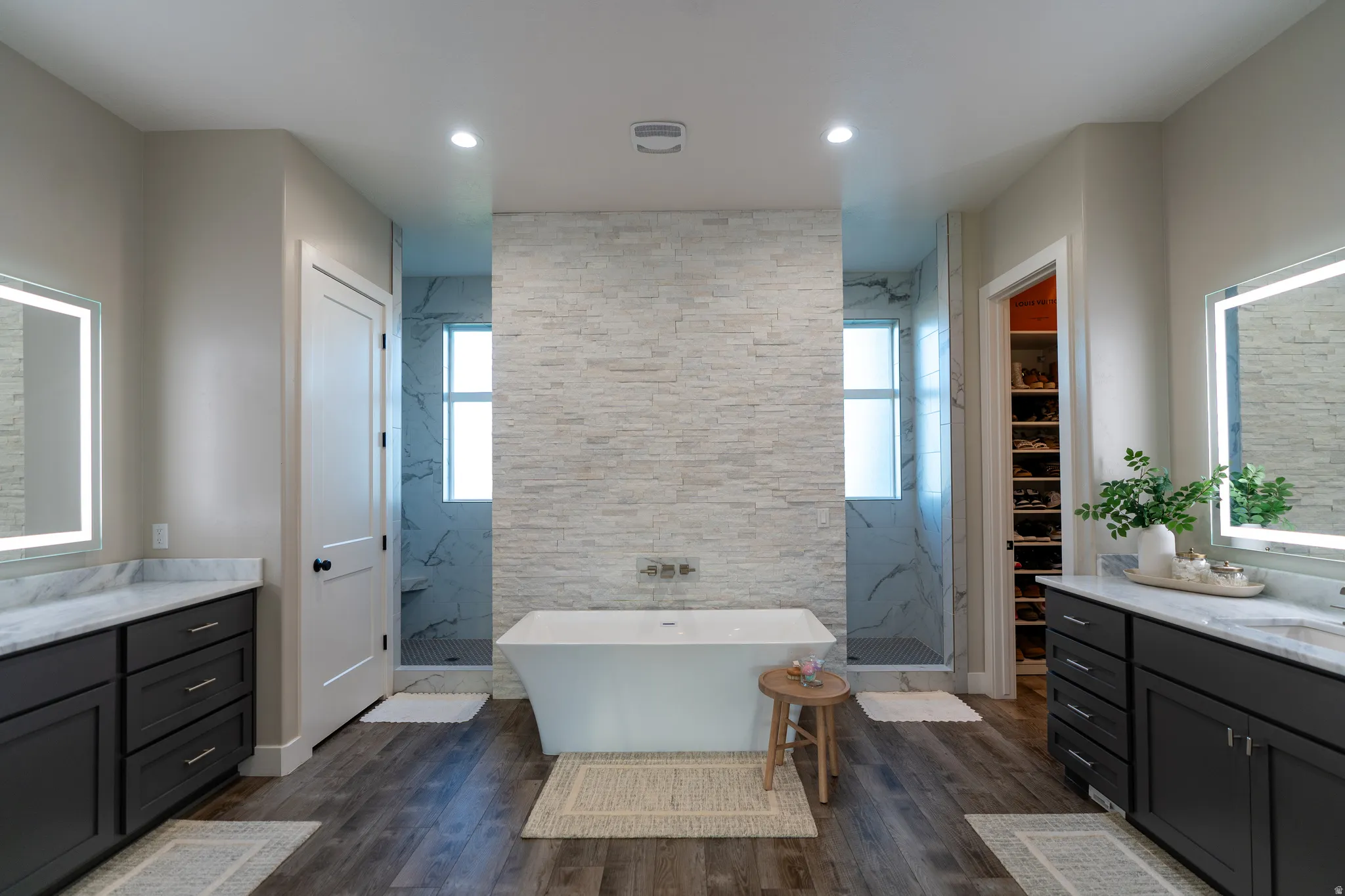 Spa-inspired bathroom featuring a freestanding soaking tub, dual glass-enclosed showers with marble-look tile, and twin vanities with illuminated mirrors