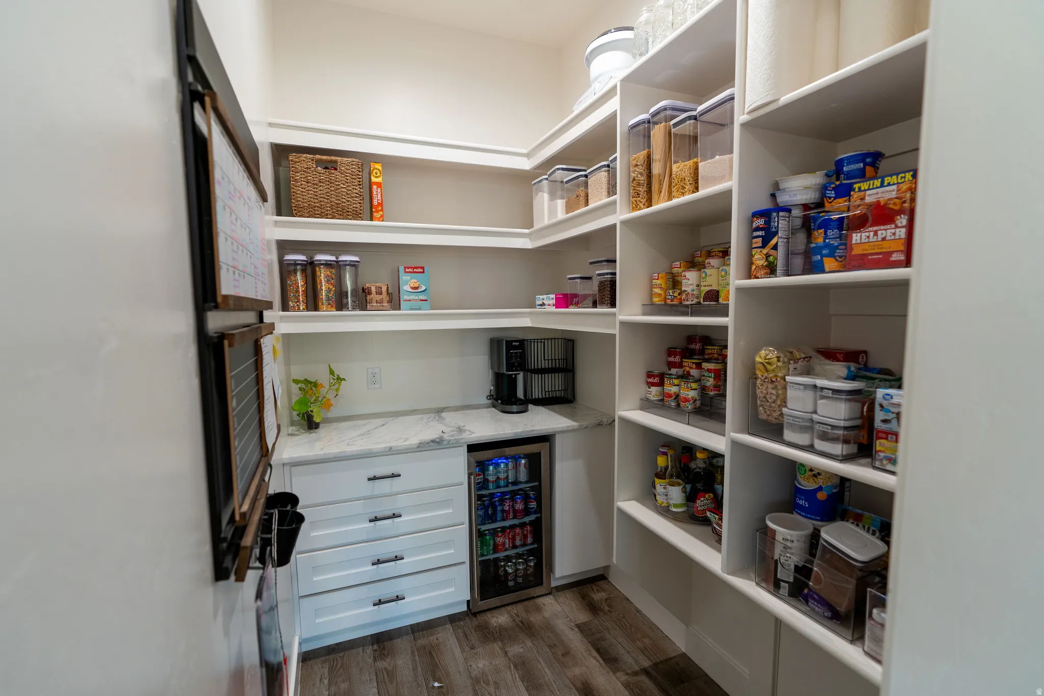 Well-appointed pantry featuring wood-finish flooring, white built-in shelving, a built-in beverage refrigerator, and a light-toned countertop
