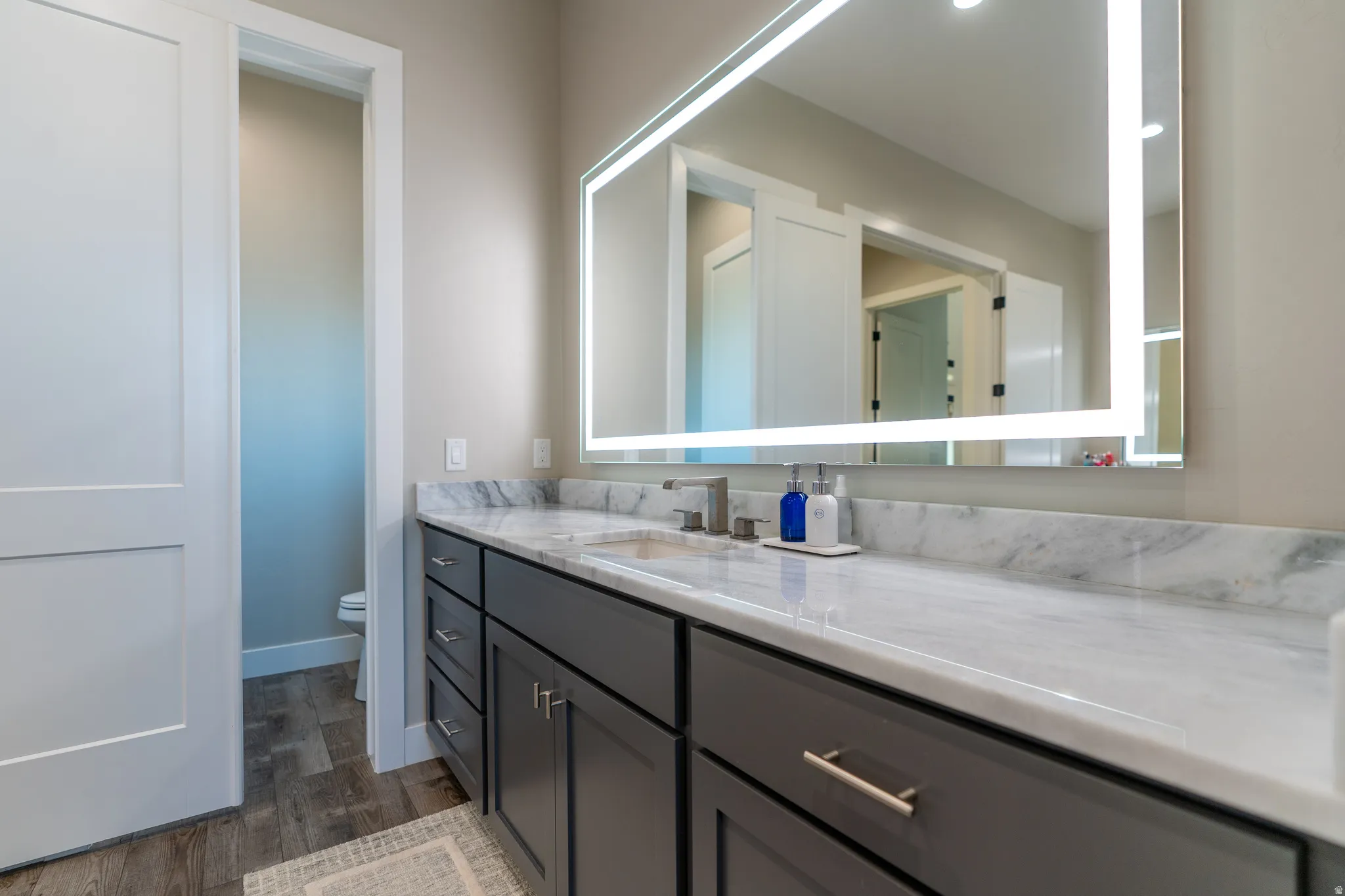 Vanity featuring a stone countertop, an integrated sink, a rectangular illuminated mirror, and dark-toned cabinetry with brushed nickel hardware