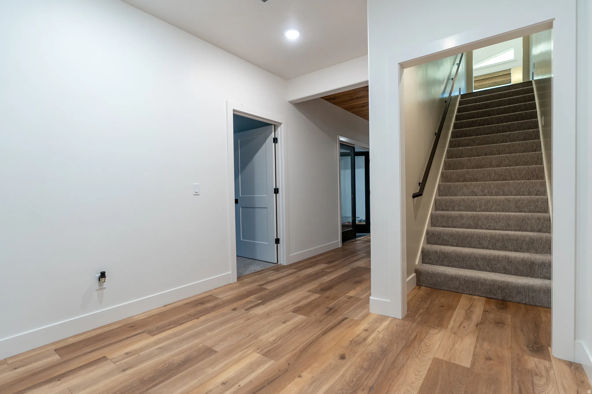 Carpeted staircase with black handrail, wood-finish flooring, white walls, and recessed lighting