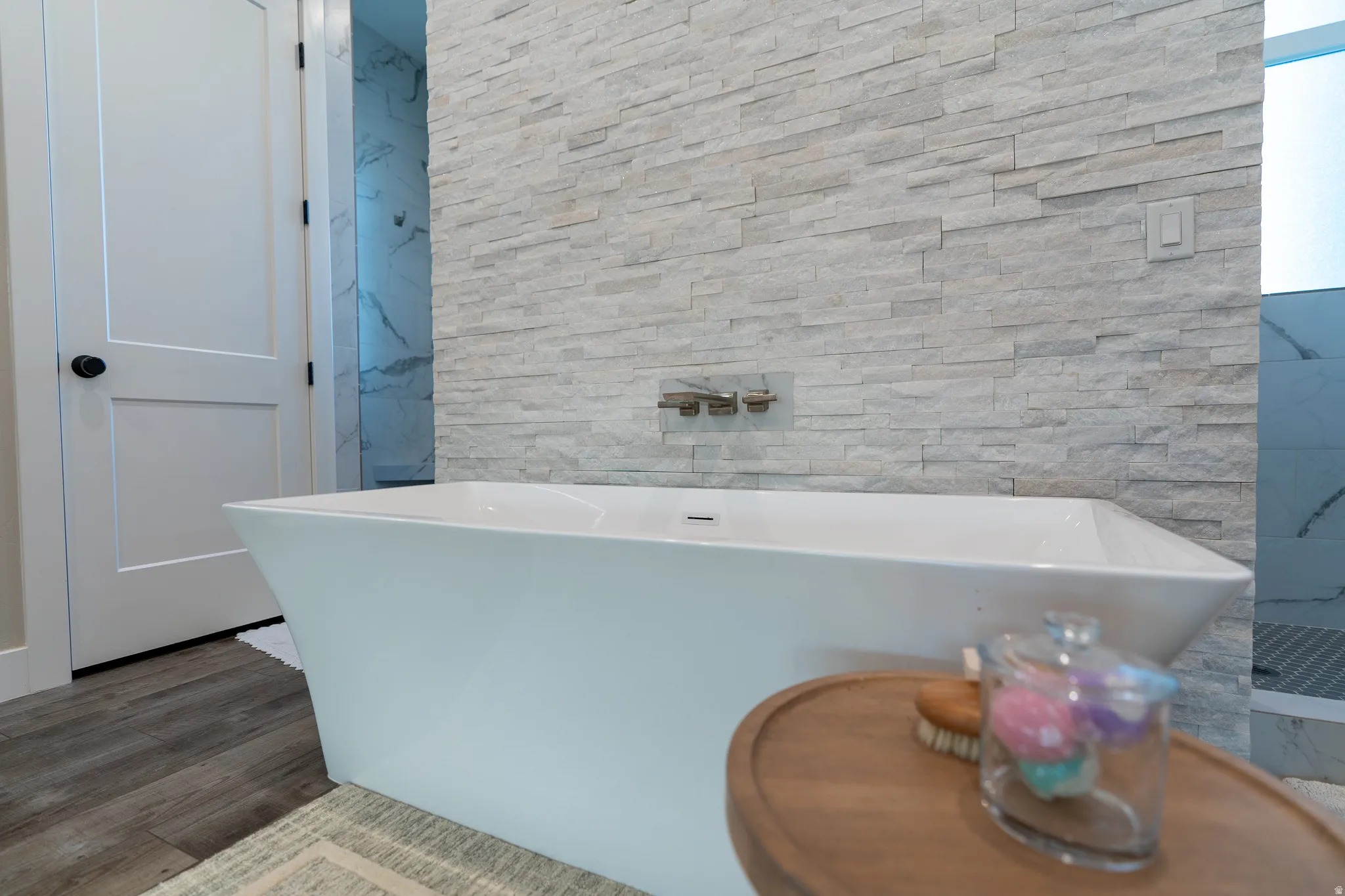 Freestanding soaking tub with wall-mounted chrome fixtures, stone-look accent wall, and wood-finish flooring