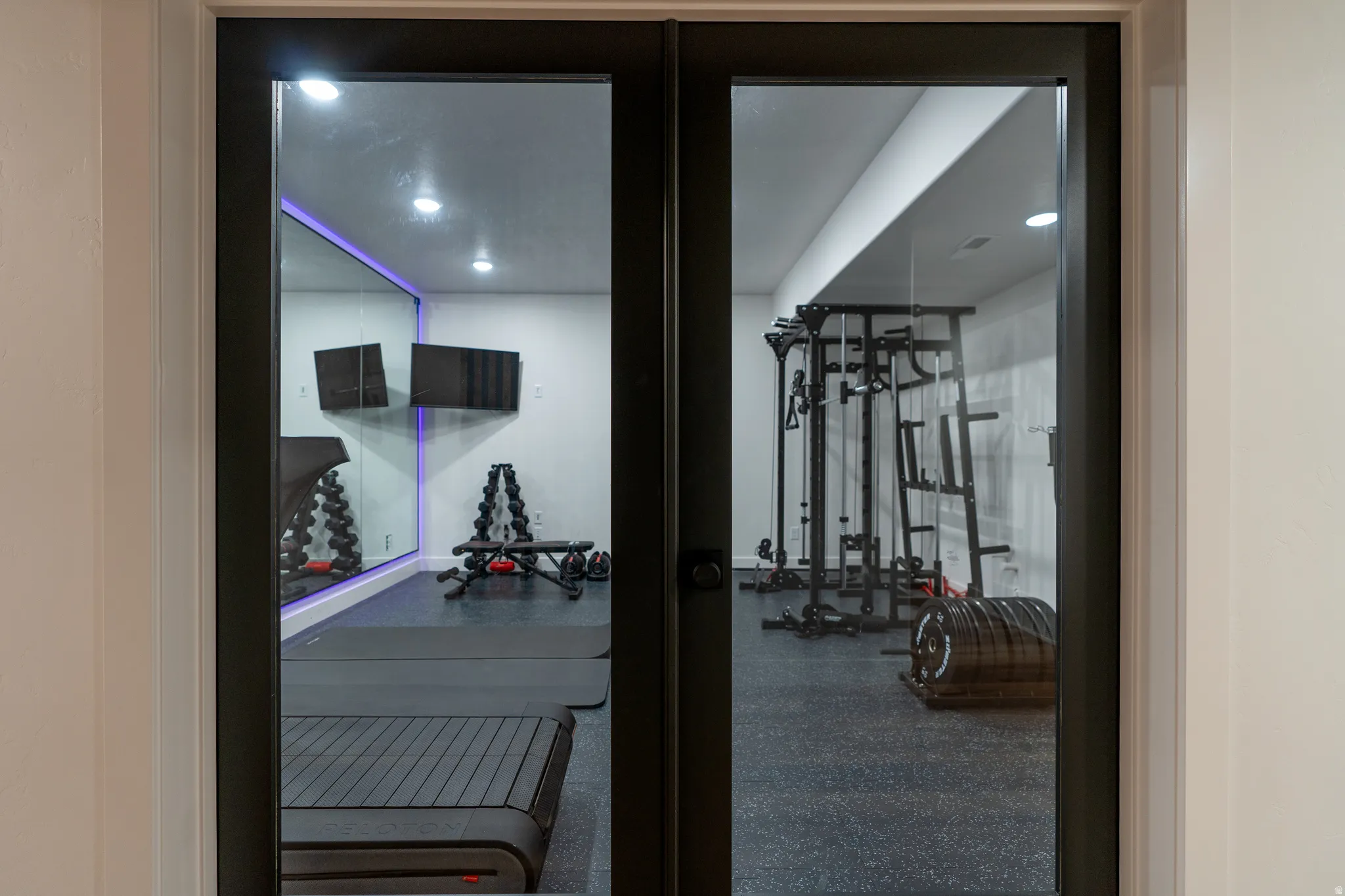 Double glass doors opening to a dedicated fitness space featuring recessed lighting, a full-wall mirror with accent lighting, and durable rubber flooring