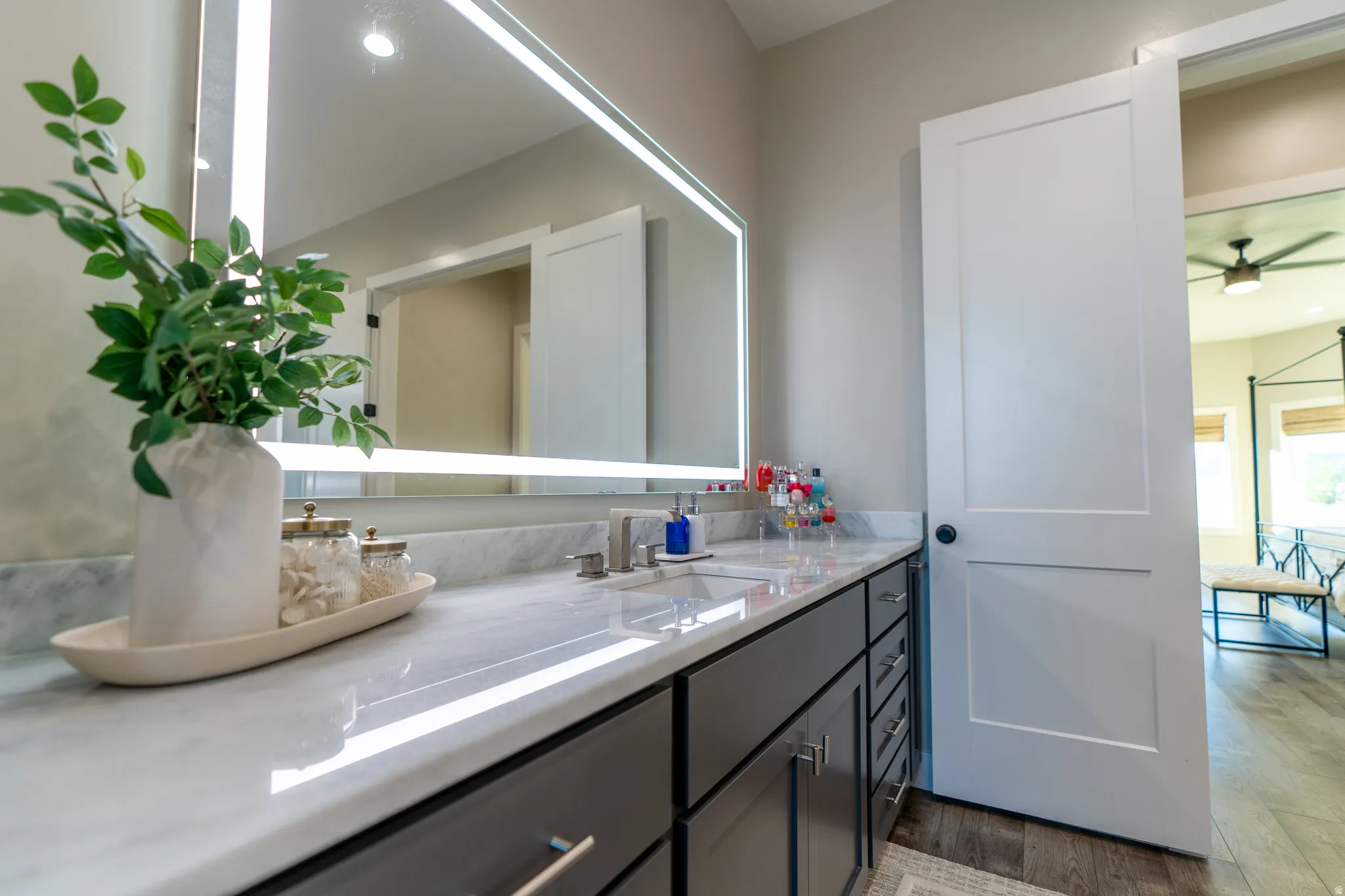 Bathroom vanity featuring a lighted mirror, dual sinks, stone countertop, dark wood-finish cabinetry, and wood-finish flooring