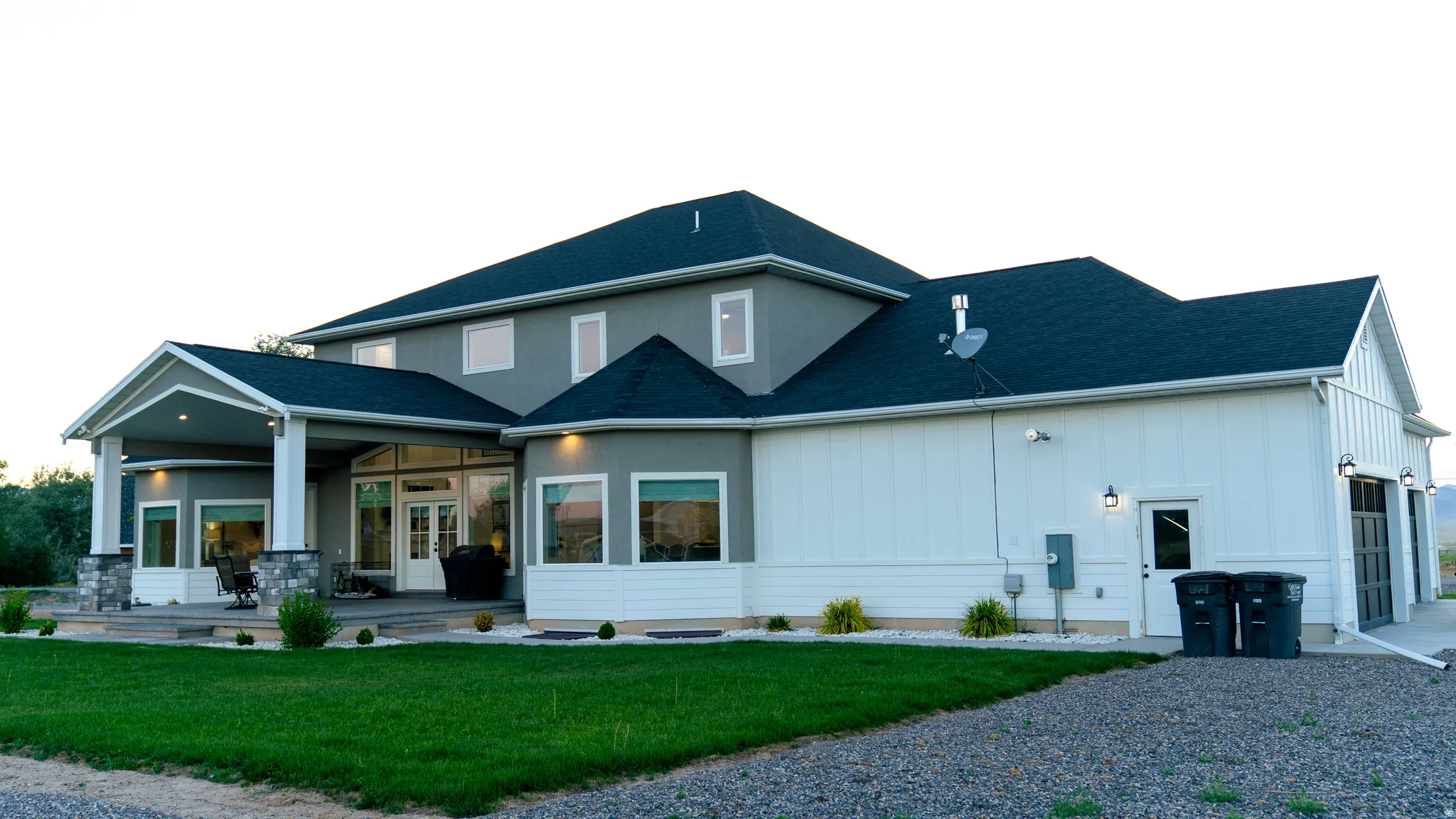 Multi-level exterior featuring a covered patio with stone-wrapped columns, a two-story stucco facade, multiple expansive windows, a large attached garage with board and batten siding, and a manicured lawn