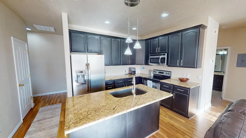 Kitchen featuring dark wood-finish cabinetry, light speckled countertops, stainless steel appliances, a center island with a double basin sink, and wood-finish flooring