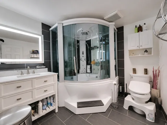 Modern bathroom featuring a contemporary multi-function shower enclosure with glass panels, a white vanity with a rectangular sink and brushed nickel faucet, an illuminated mirror, dark gray floor tiling, and a white wall-mounted cabinet