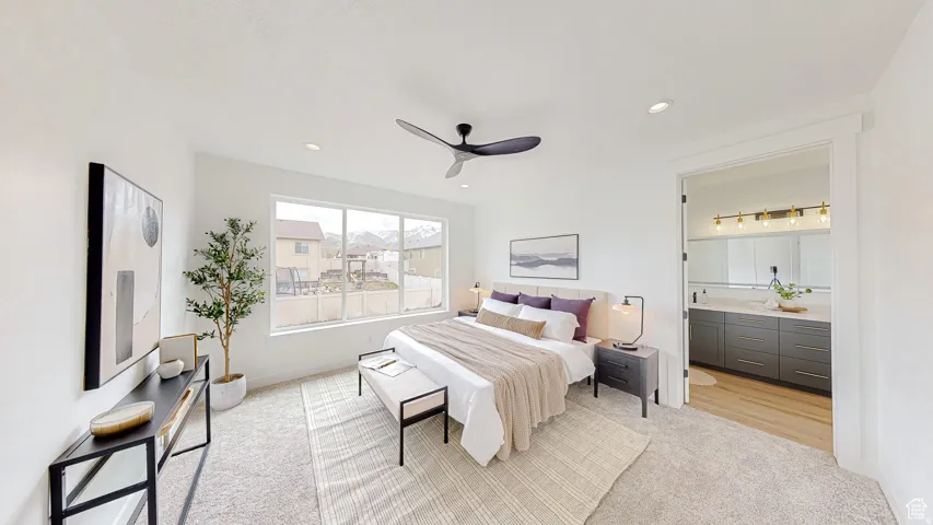 Spacious bedroom featuring light-toned carpeting, ample natural light from a large window, a modern ceiling fan, recessed lighting, and an adjacent bathroom with a double vanity and light wood-finish flooring