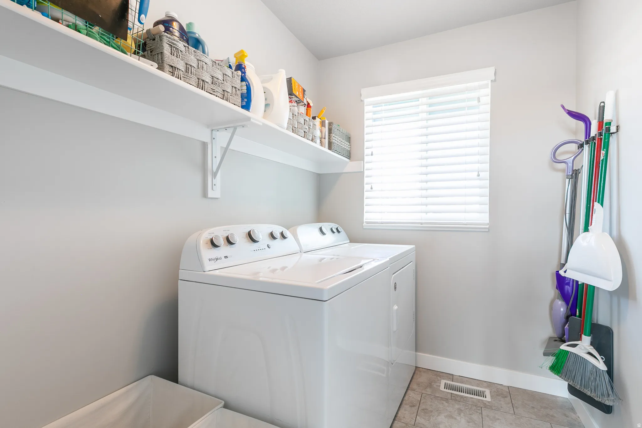 Dedicated laundry space featuring a white top-load washer and dryer set, light grey wall paint, white floating shelves, and a window with white blinds