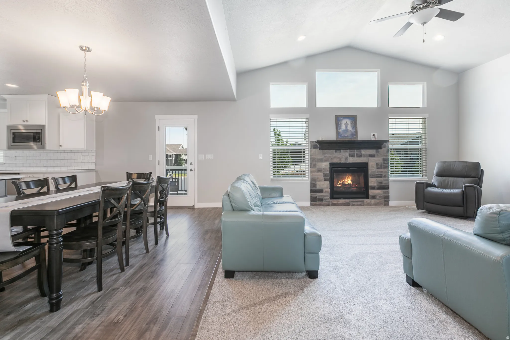 Vaulted ceiling living area featuring a stone-faced fireplace with hearth, multiple windows, and a ceiling fan