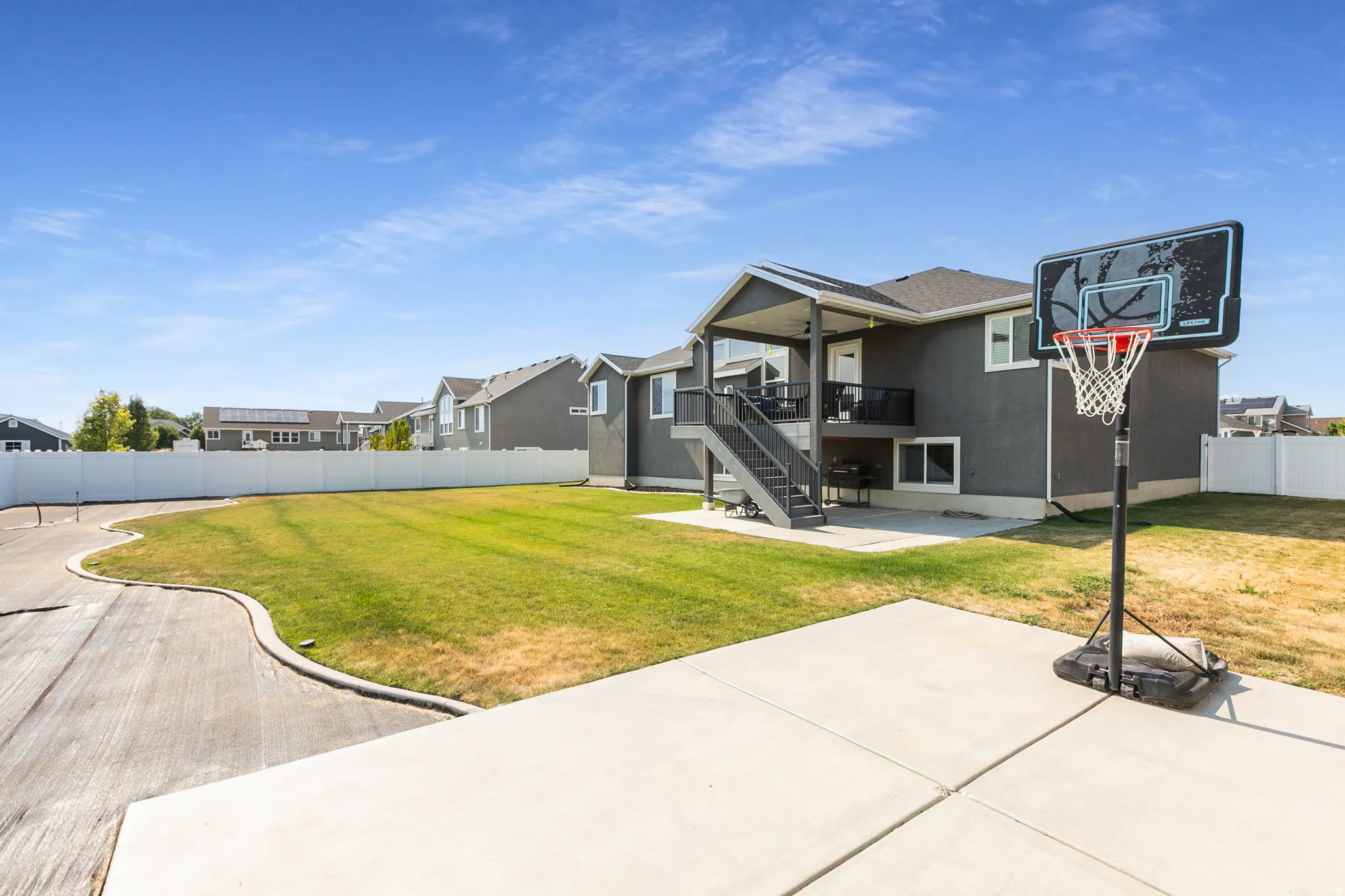 Expansive backyard featuring a large concrete patio, a covered deck with dark railings, exterior stairs, and a substantial lawn area