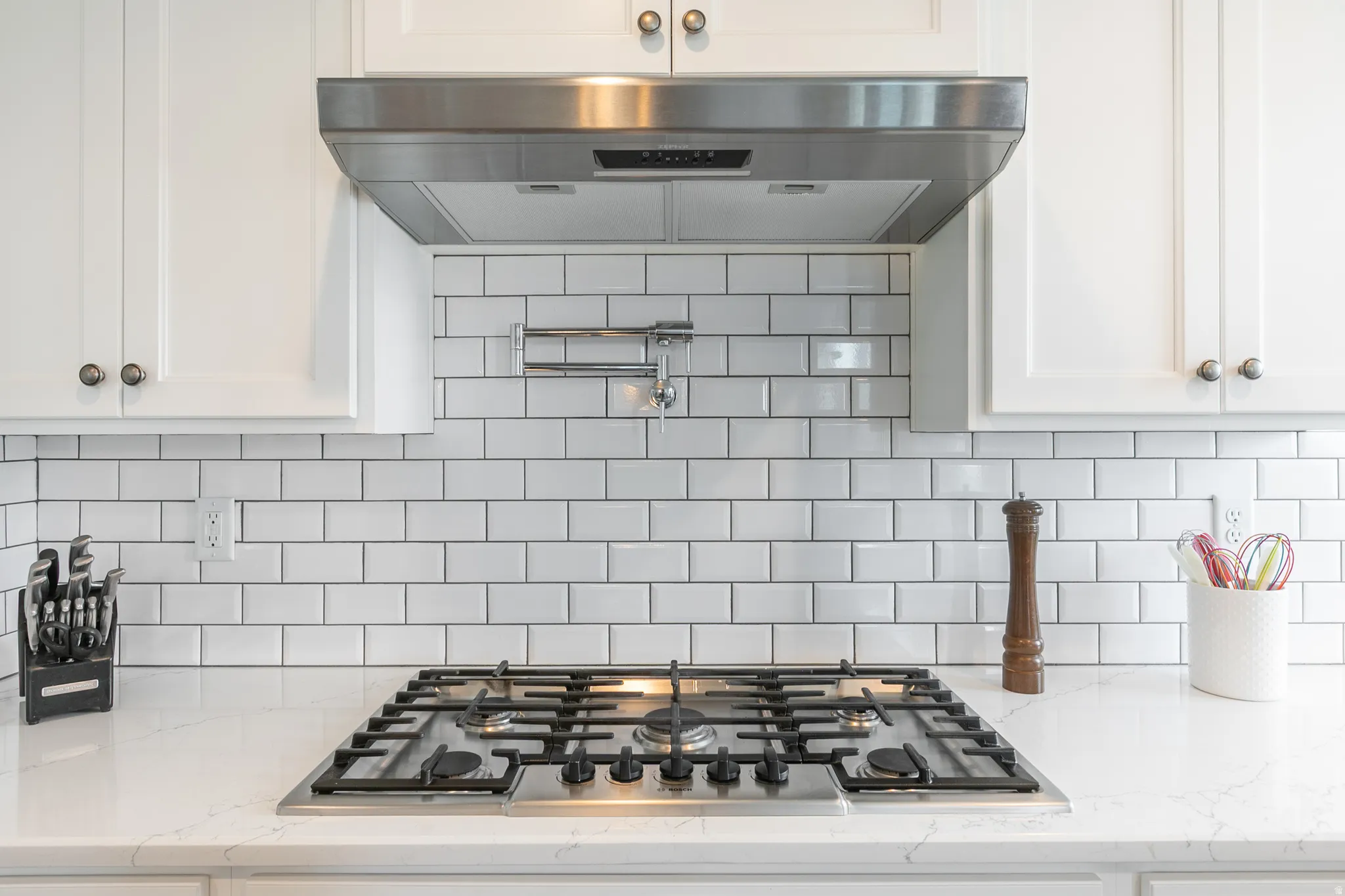Stainless steel gas range with a pot filler, set into a light-toned countertop, complemented by white subway tile backsplash and white cabinetry with brushed nickel hardware