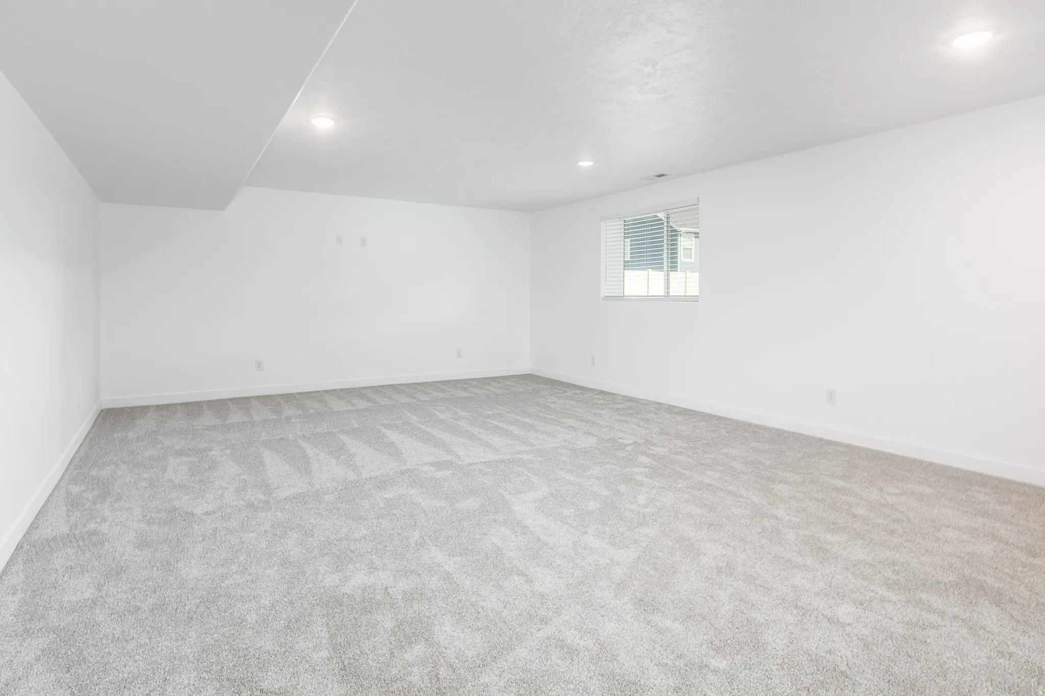 Spacious room featuring light gray carpeting, white walls, recessed lighting, and a single window with horizontal blinds