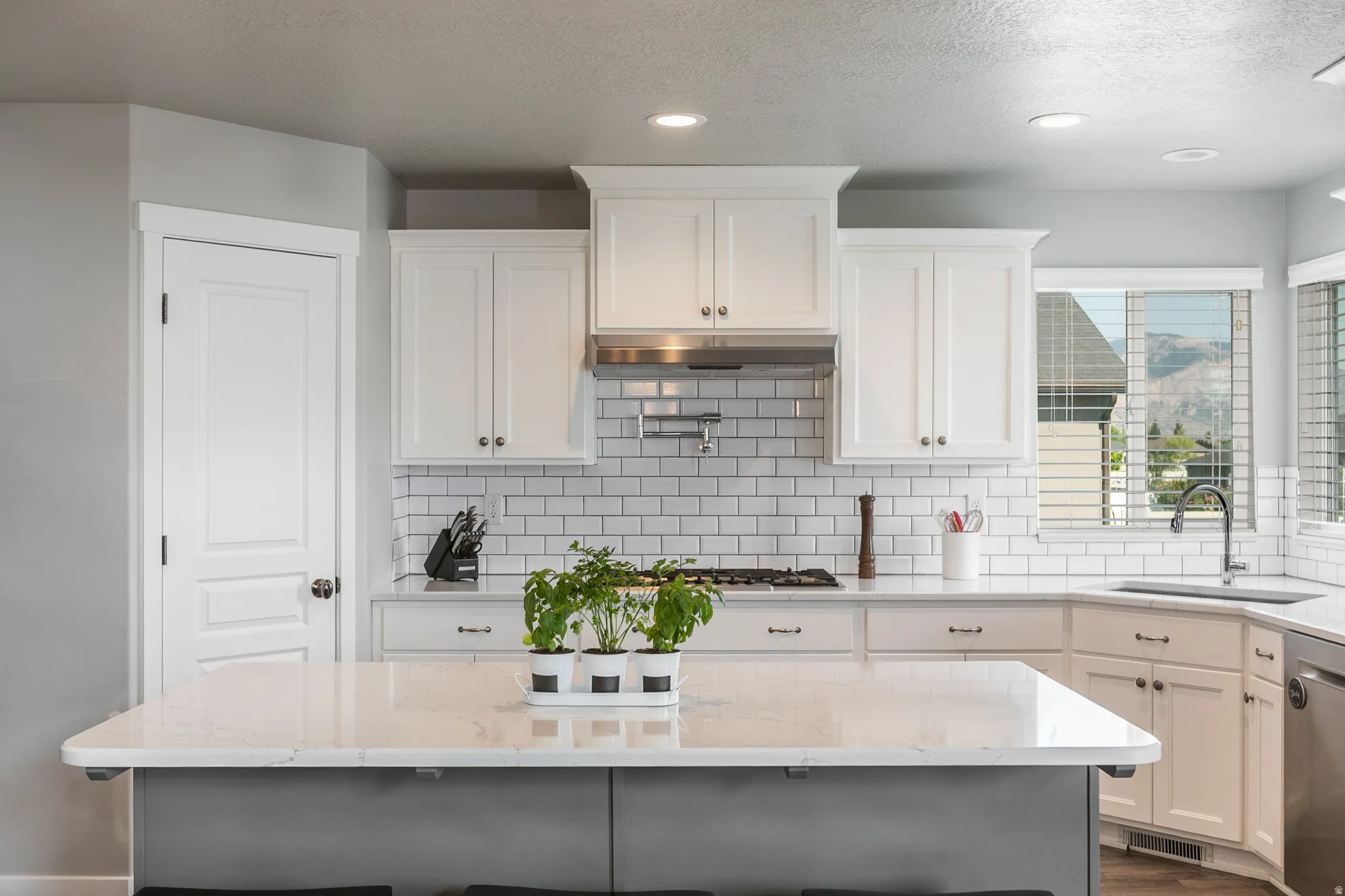 Modern kitchen featuring a large central island with a light-toned countertop, white cabinetry, subway tile backsplash, stainless steel range hood, and recessed lighting