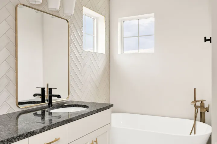 Herringbone tile wall featuring a rectangular mirror with a gold-tone frame, white vanity with black countertop, freestanding bathtub, and square windows