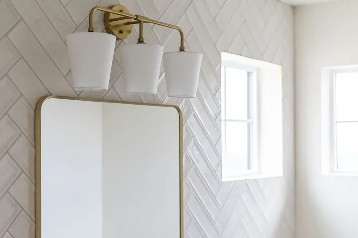 Modern bathroom featuring herringbone-pattern white tile walls, a brass-finished three-light vanity fixture with cylindrical frosted glass shades, and a brass-framed mirror with clean lines