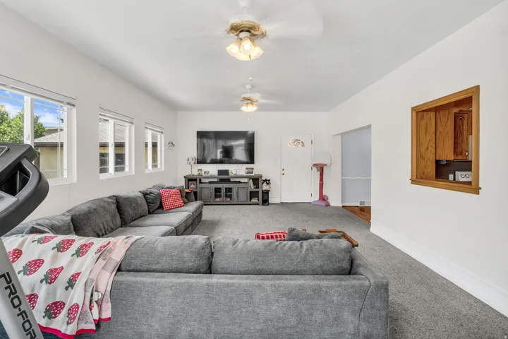 Spacious living area featuring light gray carpet flooring, multiple windows, and a built-in pass-through window with wood-finish trim