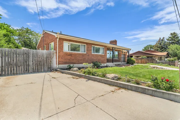 Brick exterior featuring a covered porch with dark support columns, a concrete driveway, a wooden privacy fence, and a front lawn with established landscaping