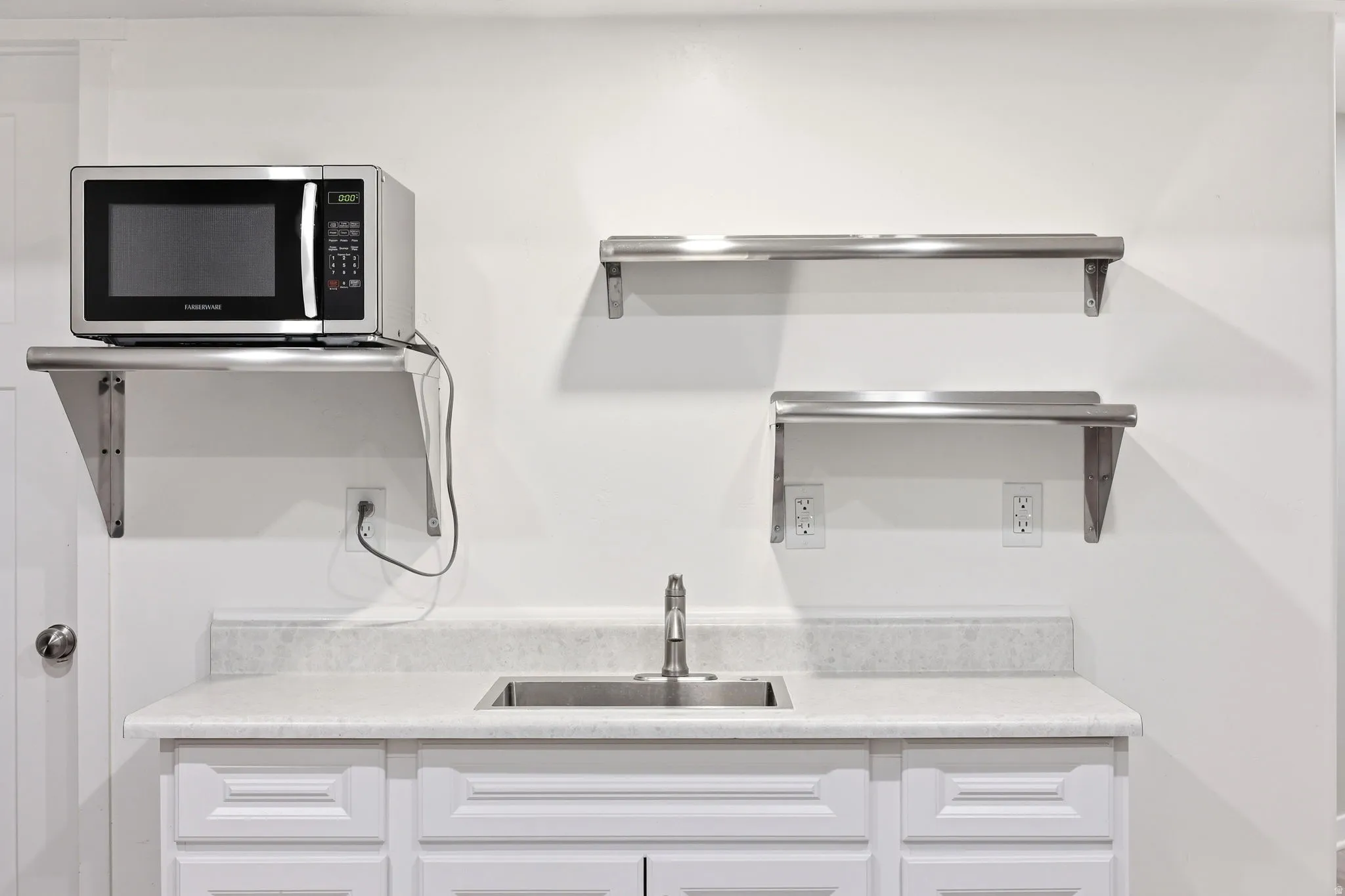 Kitchenette featuring a stainless steel sink, white cabinetry with recessed panel doors, and a light-toned countertop with a backsplash
