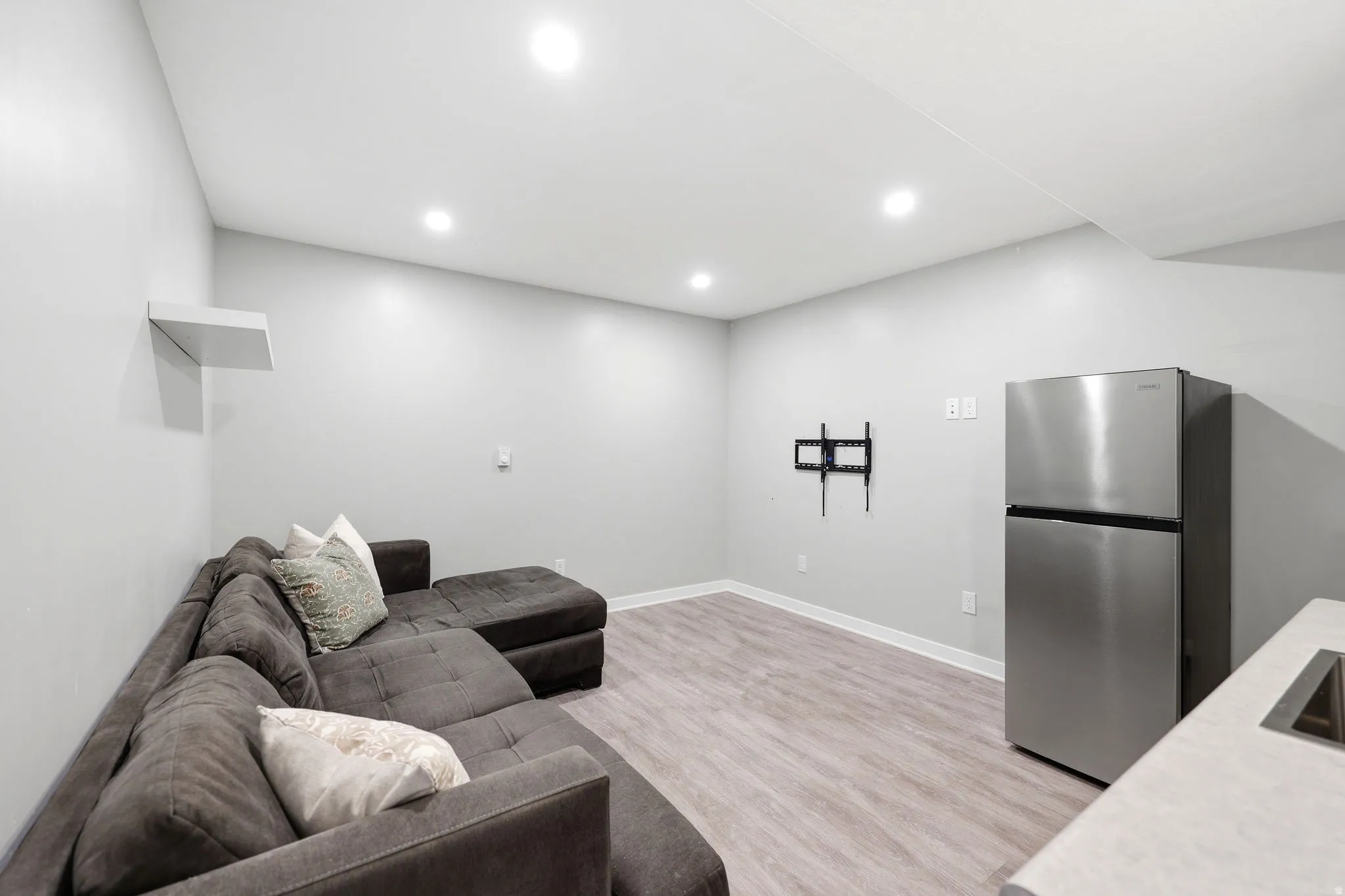 Open-concept room featuring light gray walls, recessed lighting, wood-finish flooring, a stainless steel refrigerator, and a built-in wall shelf