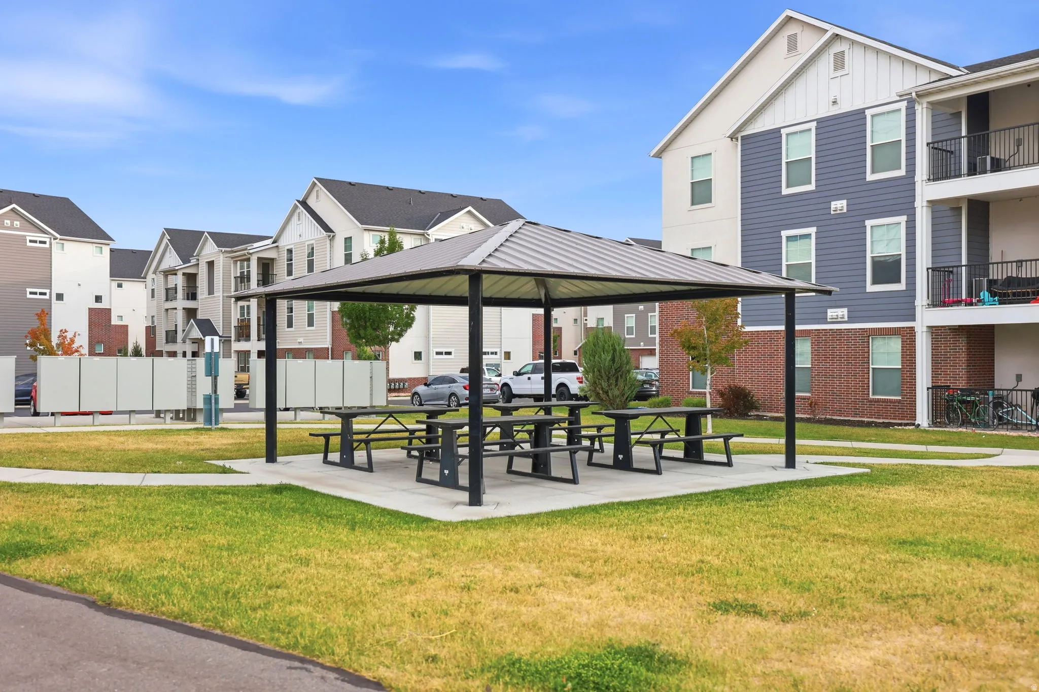 Covered outdoor seating area with multiple picnic tables, surrounded by green space, and bordered by a paved path