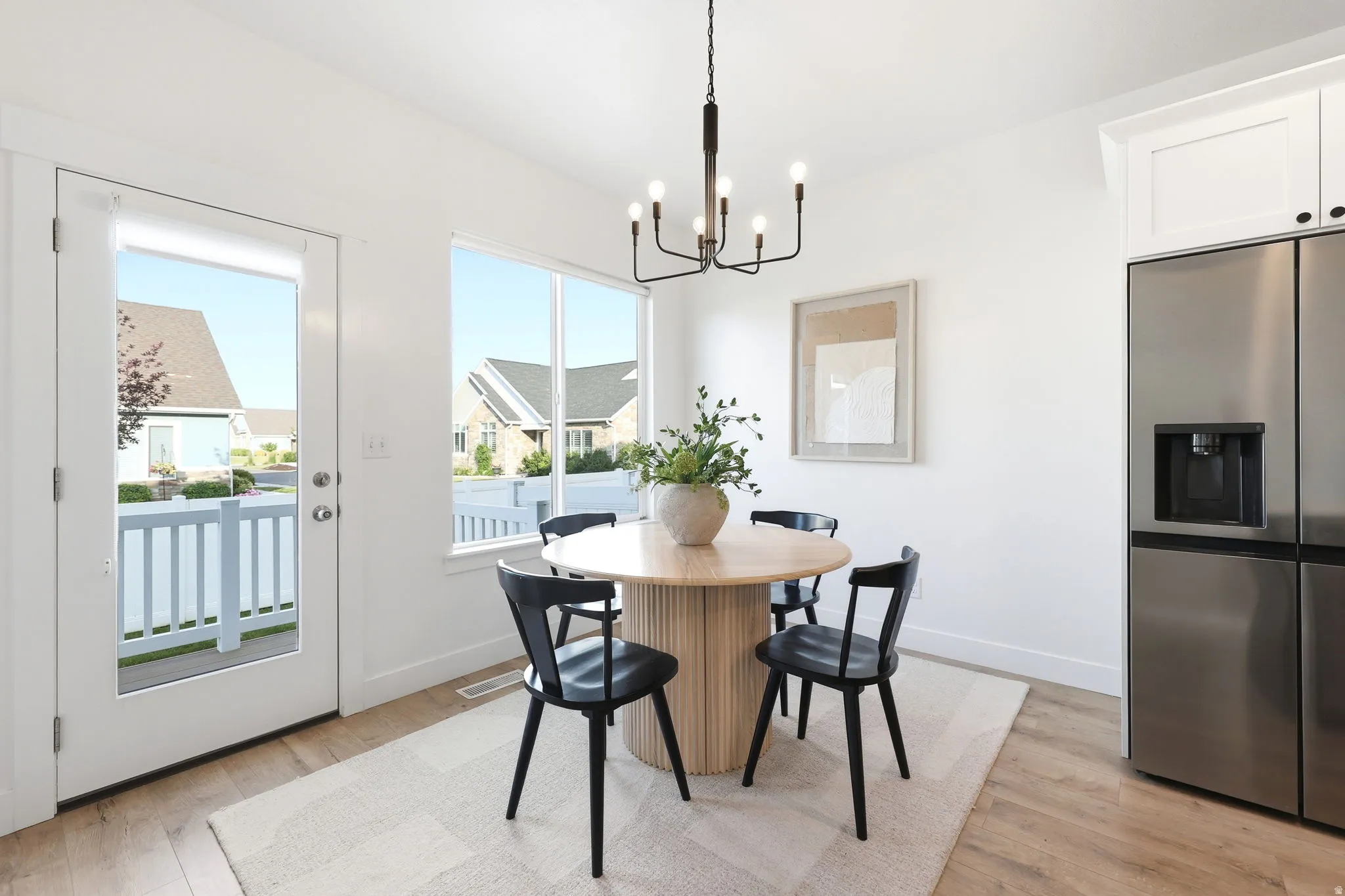 Bright dining area featuring wood-finish flooring, a contemporary black chandelier, and a stainless steel refrigerator with a built-in dispenser