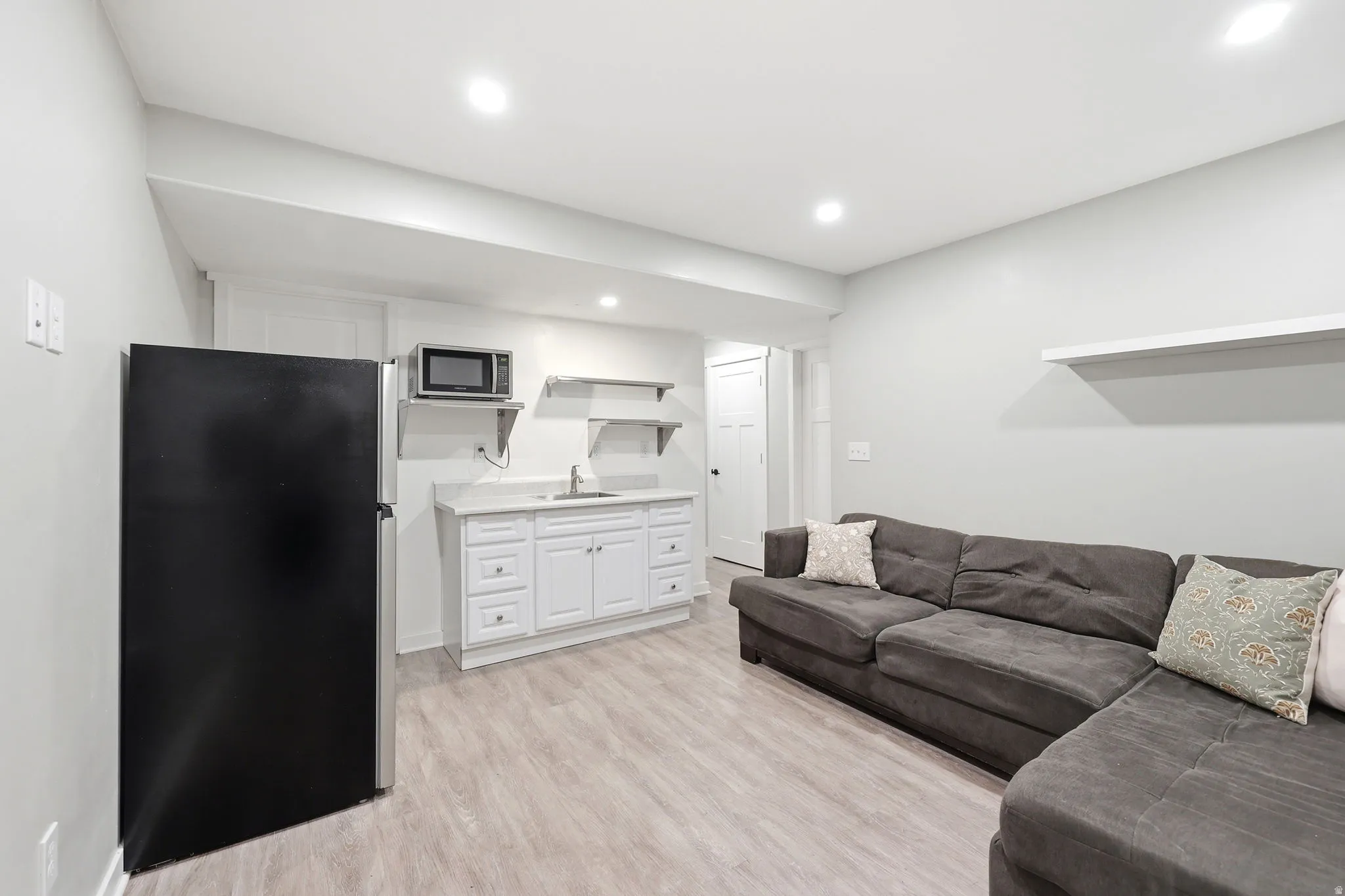 Open-concept living area featuring light-toned wood-finish flooring, a built-in kitchenette with white cabinetry, a single basin sink, and a black refrigerator