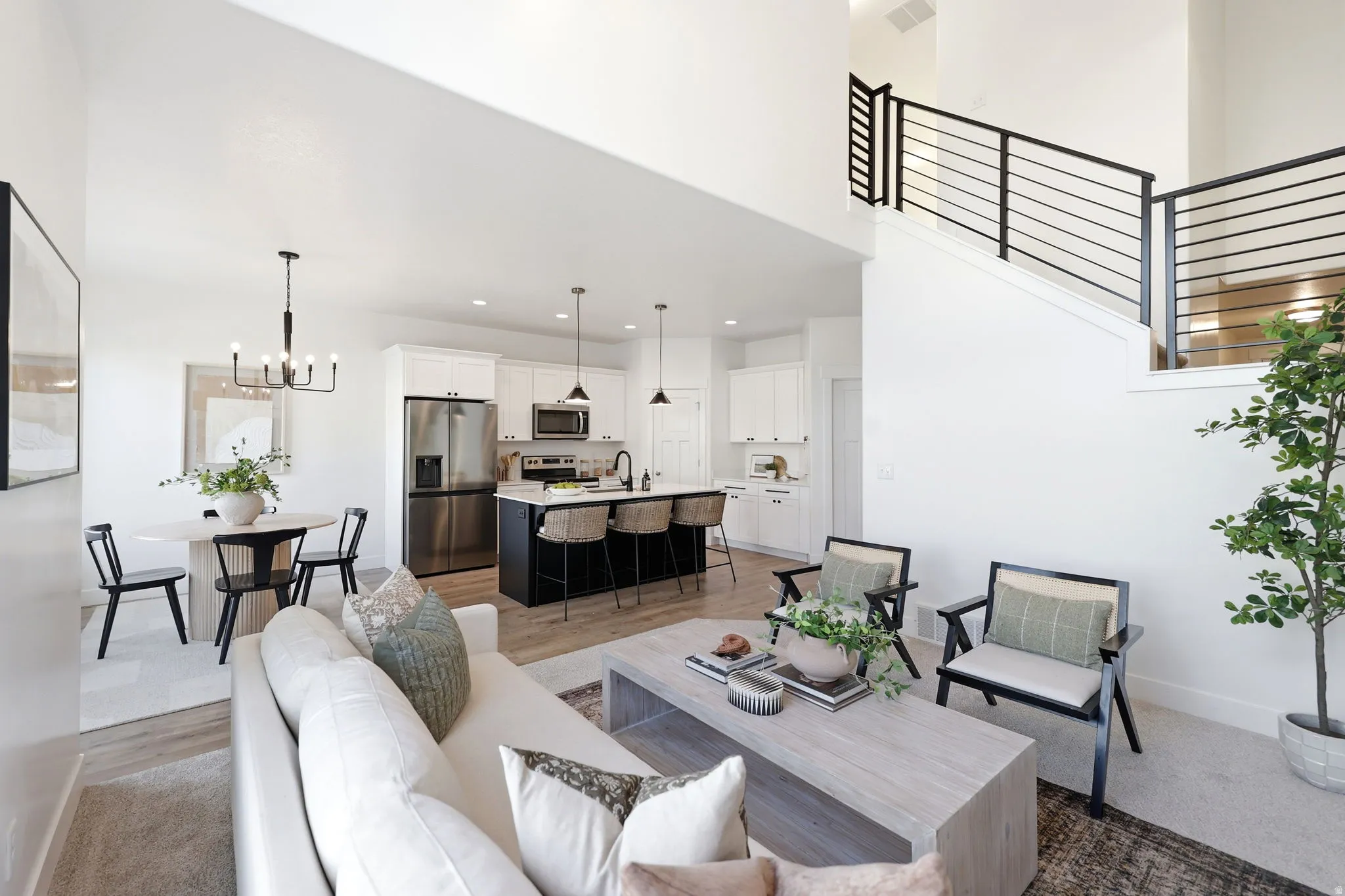 Open-concept living space featuring high ceilings, contemporary kitchen with stainless steel appliances, central island with seating, a dedicated dining area, and modern staircase railing