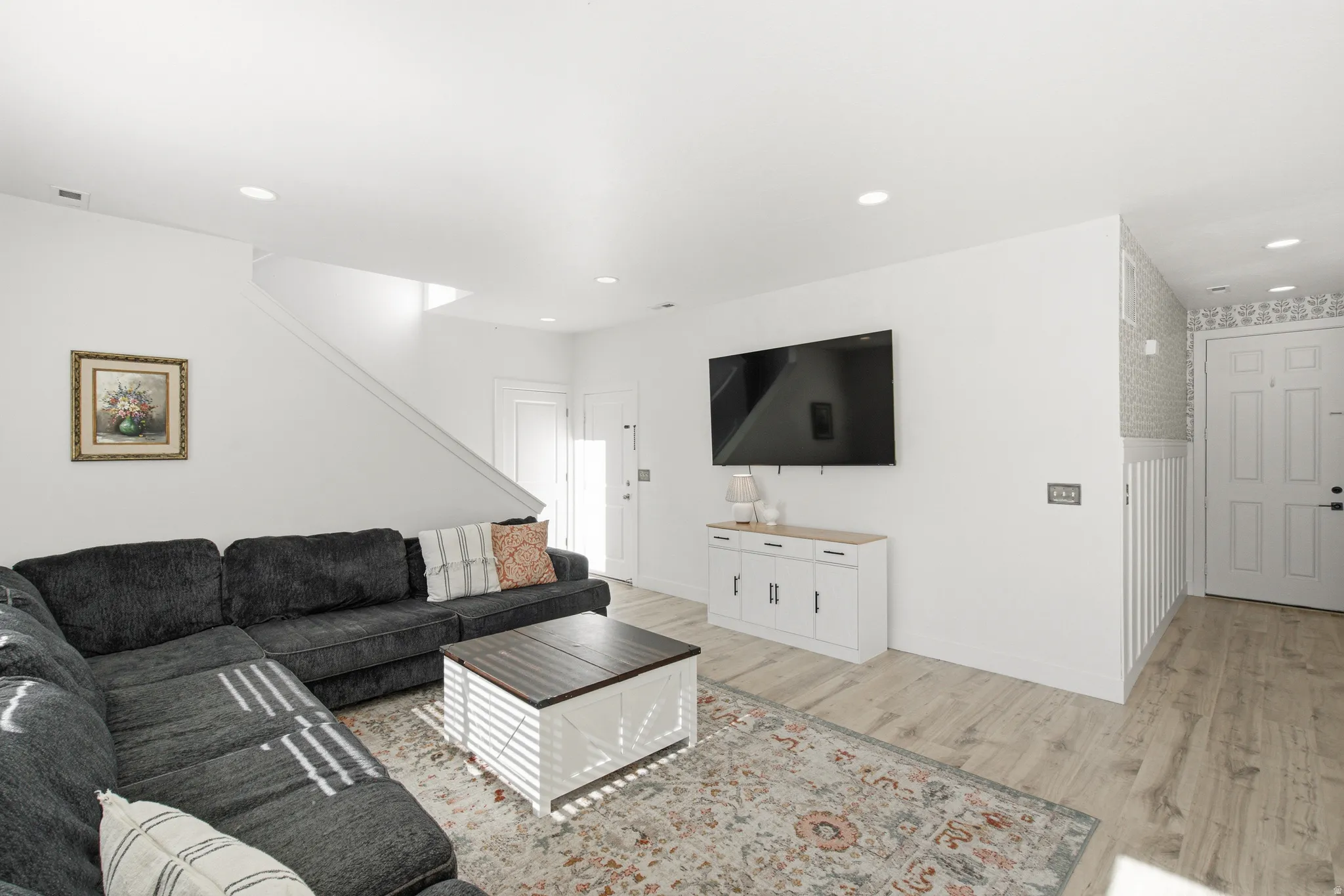 Spacious living area featuring light wood-finish flooring, recessed lighting, white walls, and a staircase with white risers
