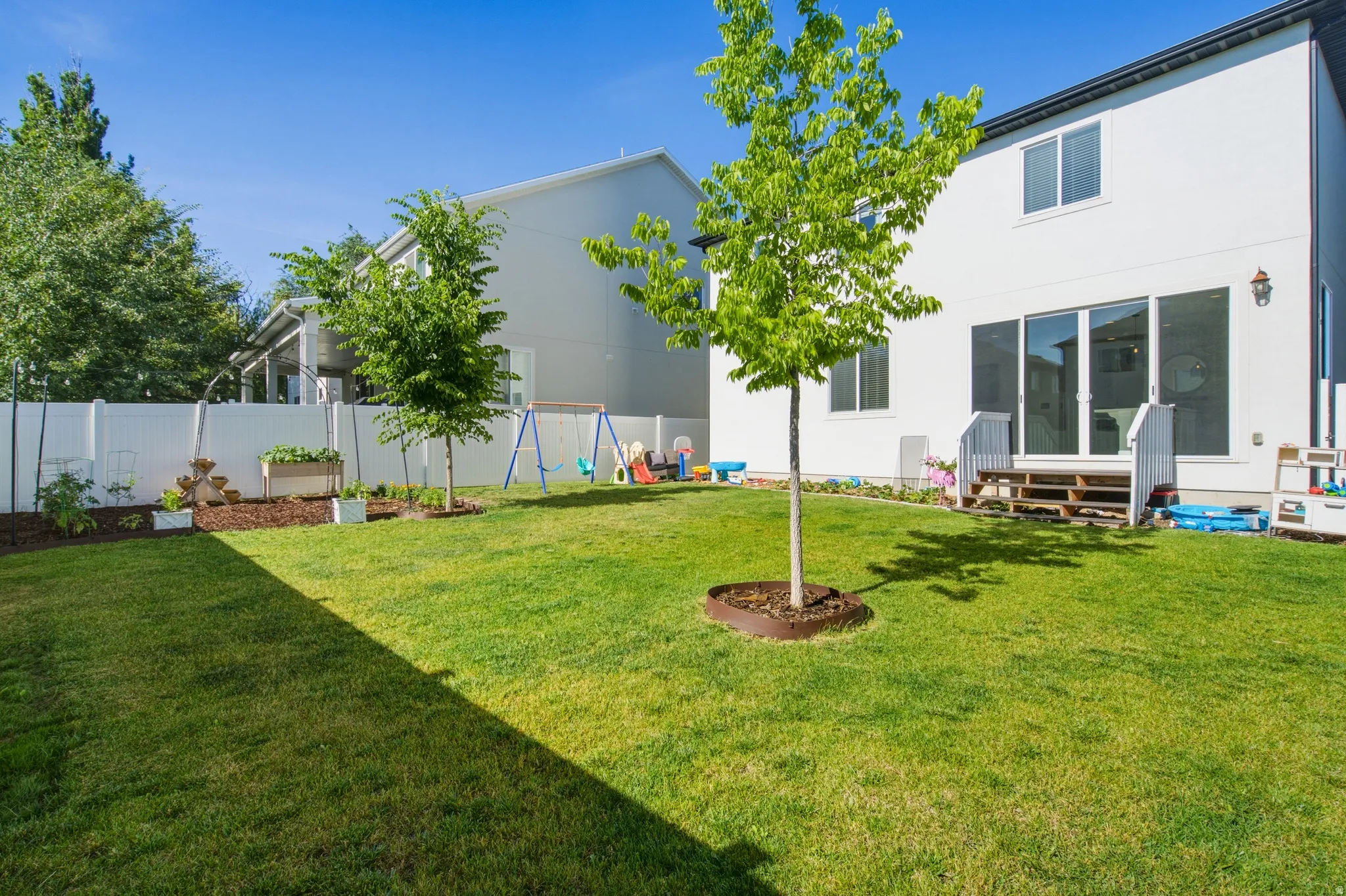 Expansive grassy backyard featuring a white privacy fence, mature trees, a raised garden bed, and a ground-level deck with steps leading to sliding glass doors