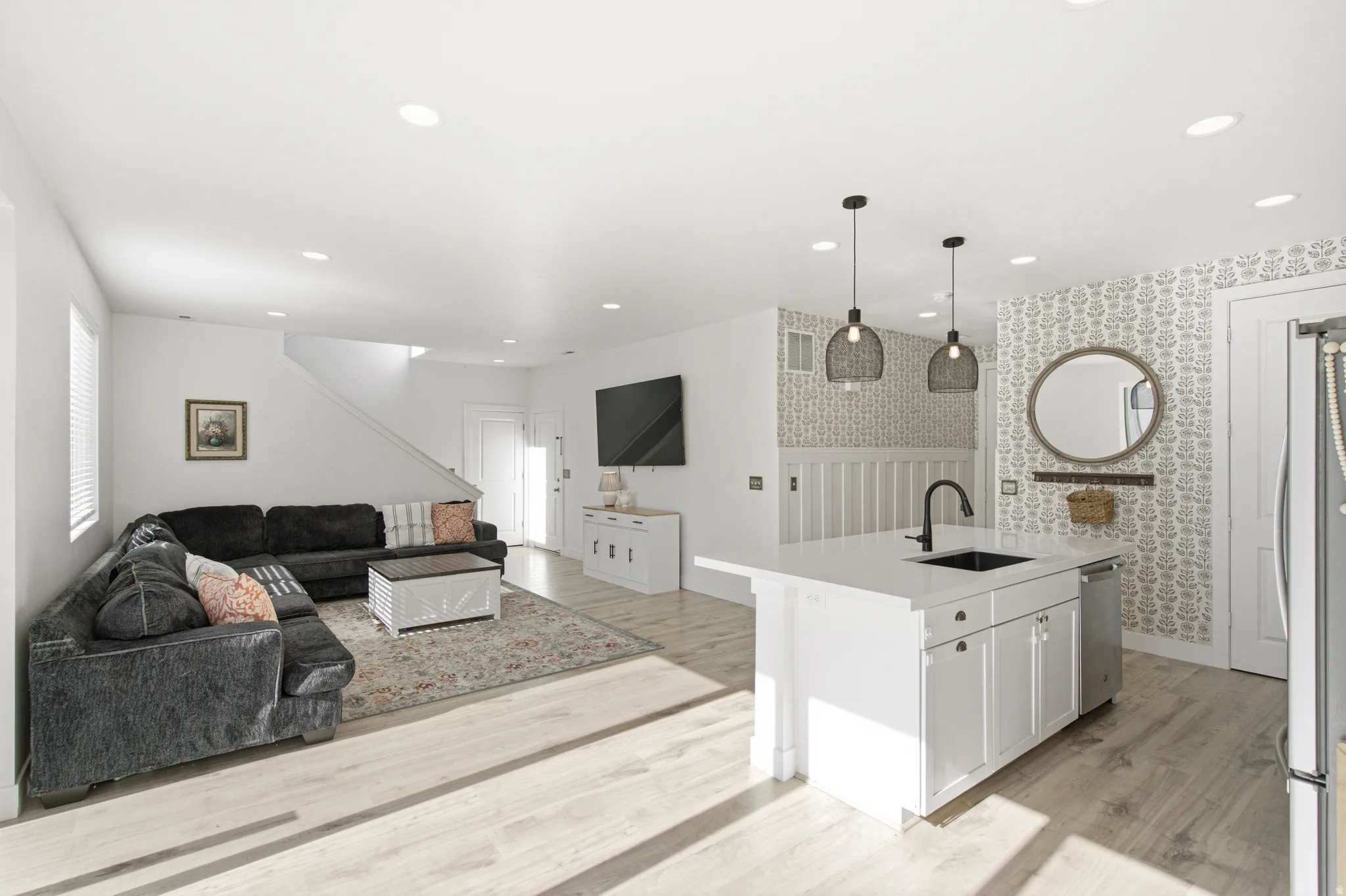 Spacious open-concept living area featuring light wood-finish flooring, recessed lighting, and a white kitchen island with a black faucet