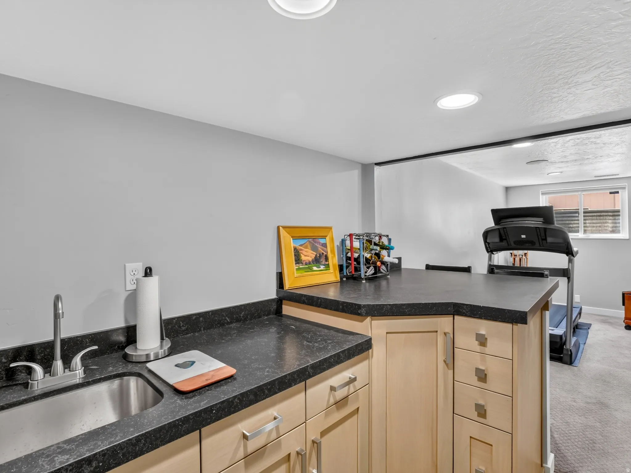 Built-in wet bar with a stainless steel undermount sink, dark countertops, light wood-finish cabinetry, and recessed lighting