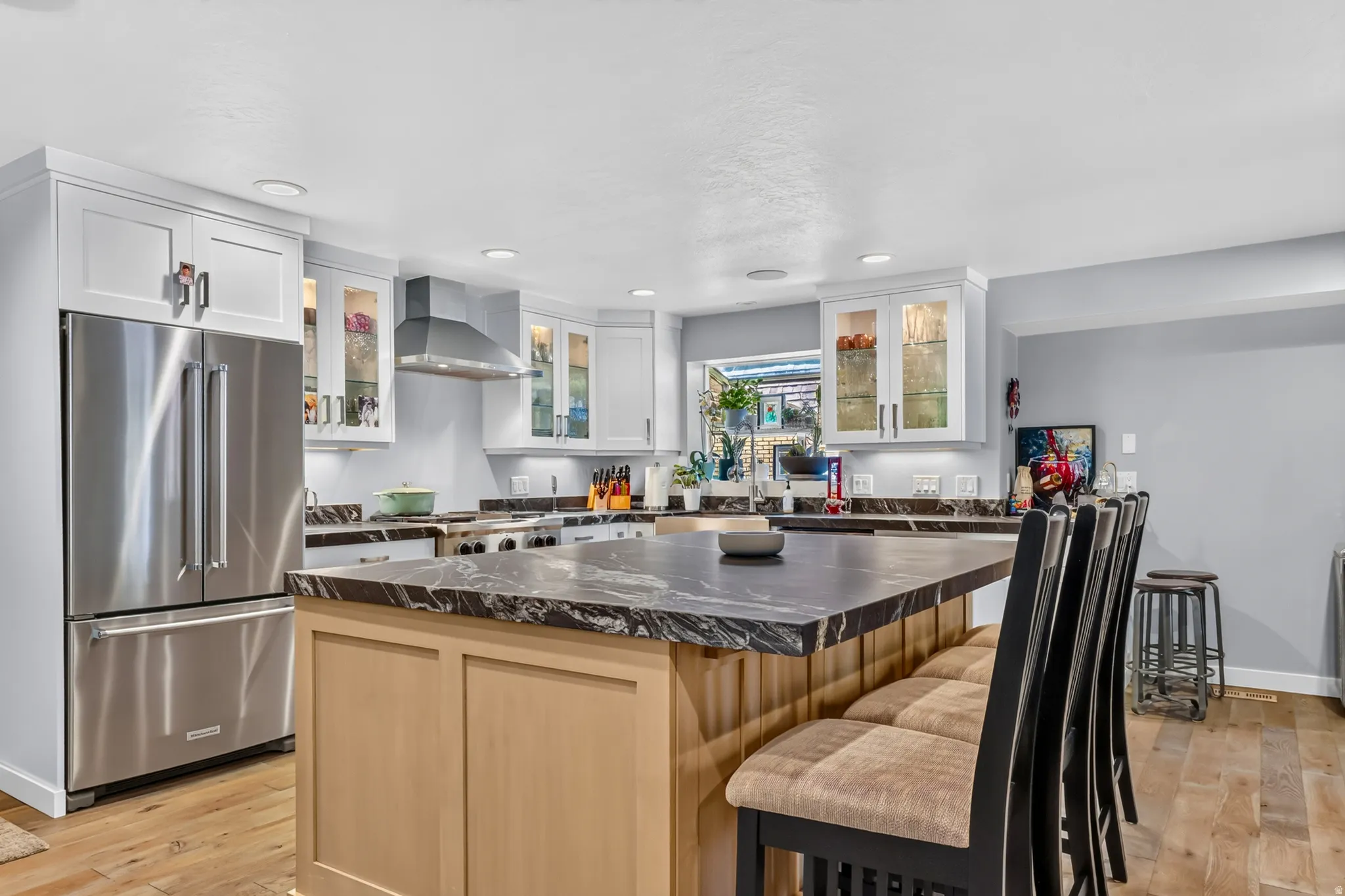 Open-concept kitchen featuring an expansive central island with seating, dark stone countertops, wood-finish flooring, white shaker cabinetry, and stainless steel appliances