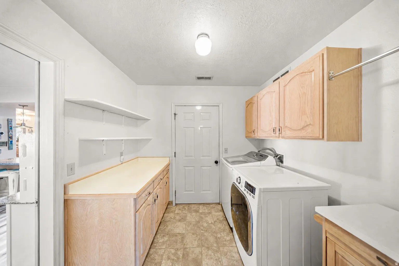 Dedicated utility room featuring light wood-finish cabinetry, a long countertop, wall-mounted shelving, overhead storage, and tile-look flooring