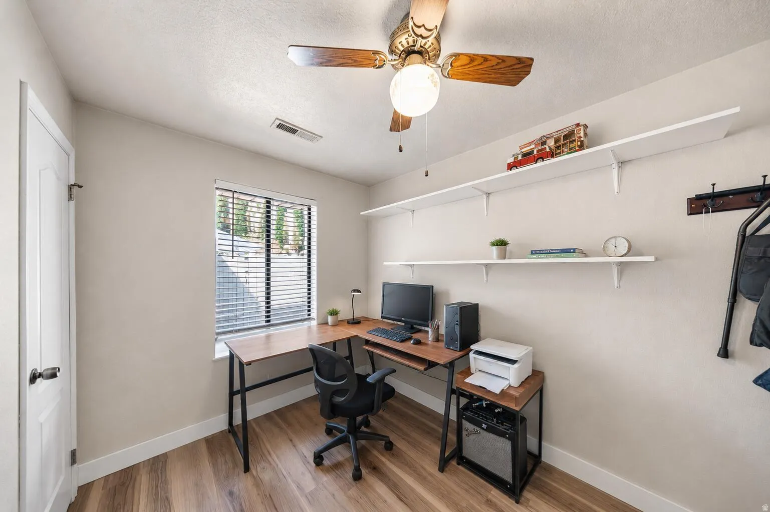 Room featuring wood-finish flooring, a window with horizontal blinds, a white door, a ceiling fan with light, and two white wall-mounted shelves